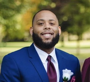 A young man with a beard wearing a blue suit, white shirt, and maroon tie, smiling outdoors with green trees in the background.