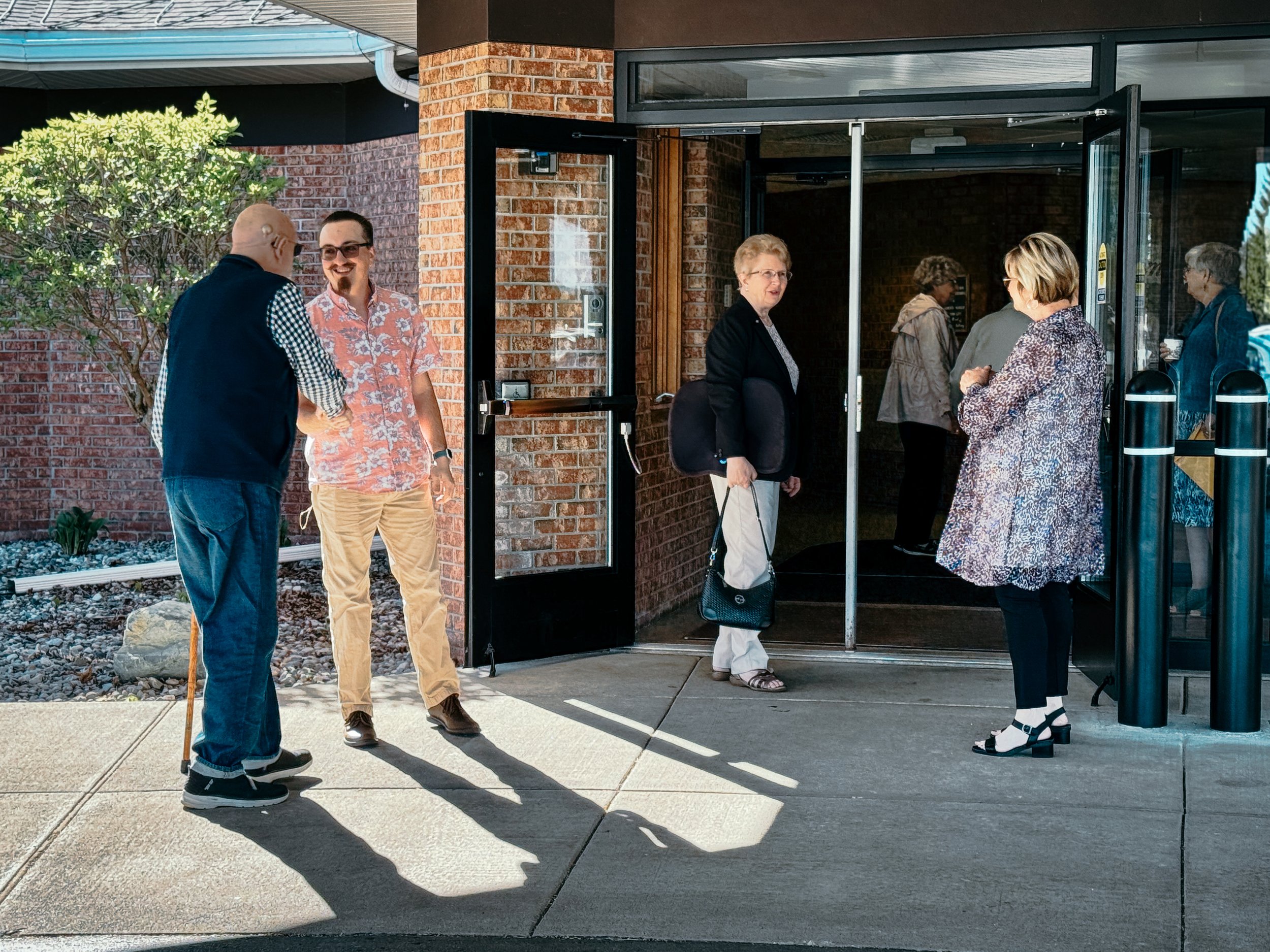A group of six people standing outside near a glass door, engaged in conversation, with a brick wall and a bush in the background.