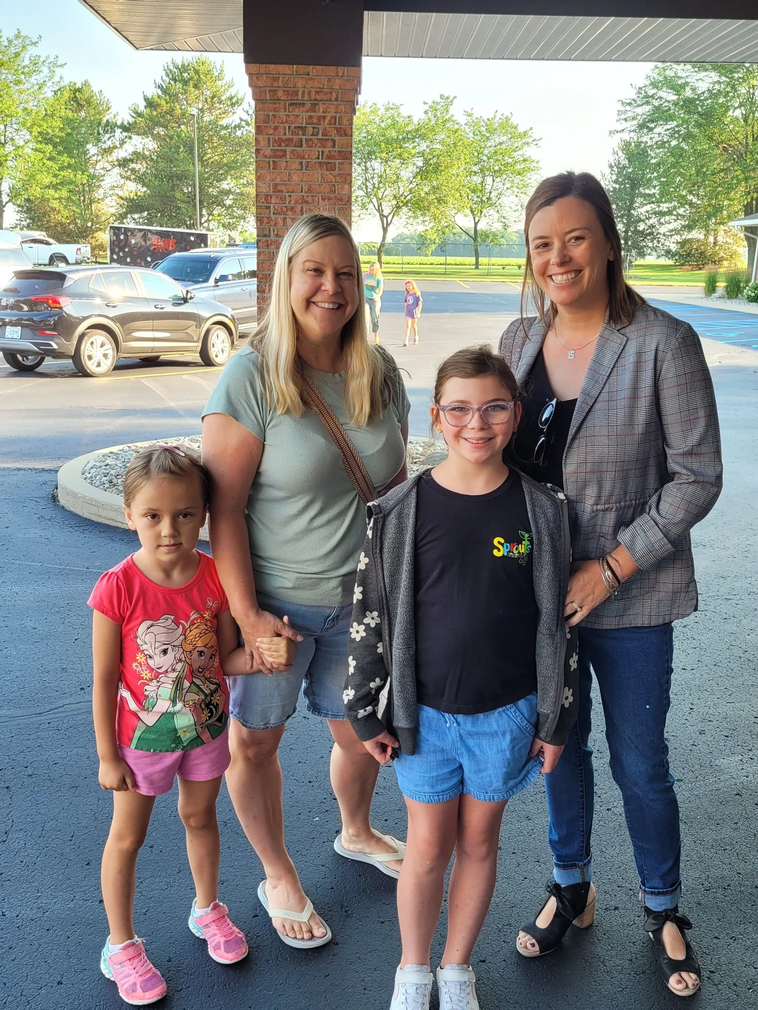 Five females, two children and three adults, are outside on a sunny day, smiling and posing for a photo. They are standing on a dark-colored parking lot with cars and trees in the background.
