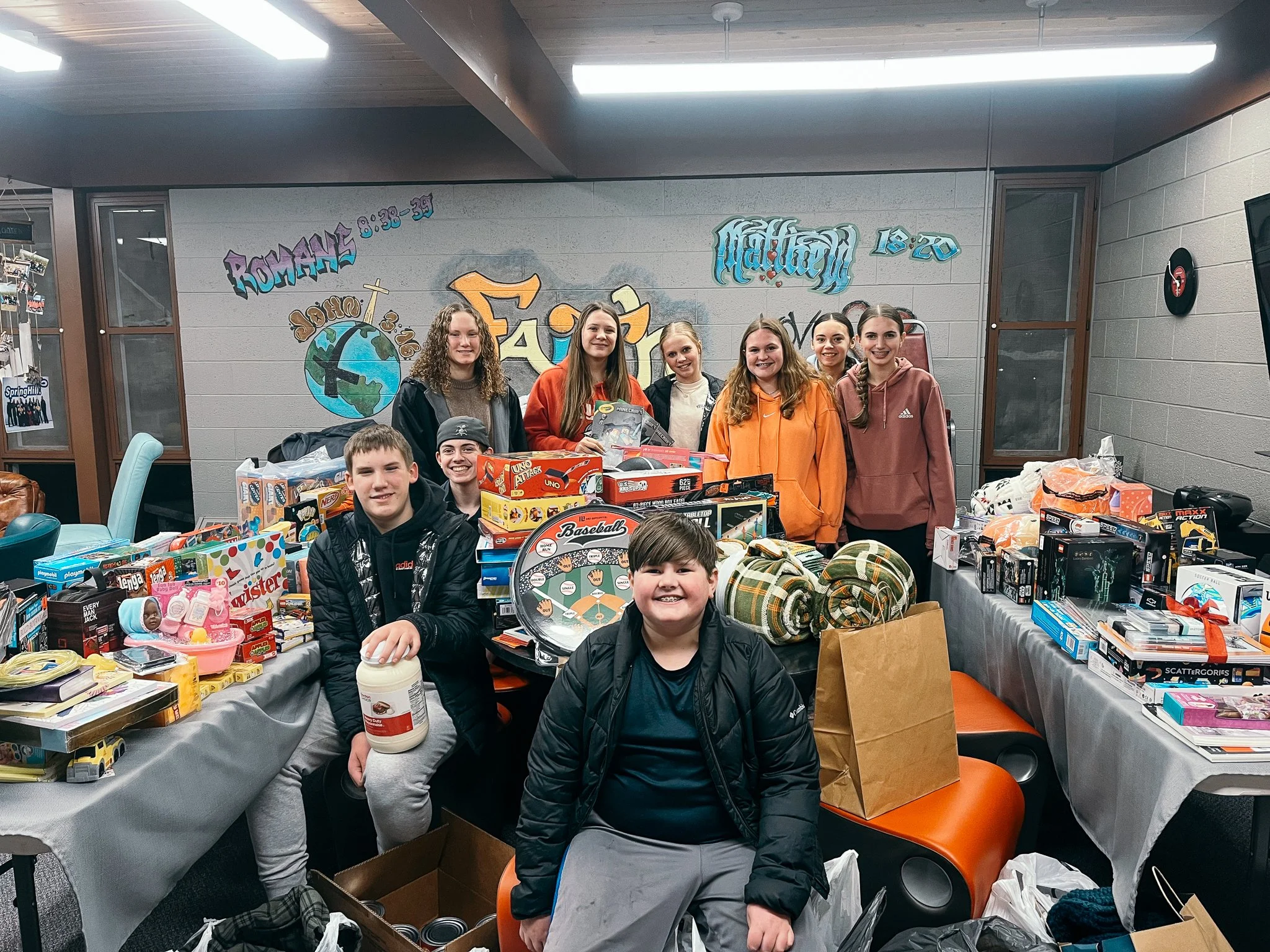 Group of young boys and girls standing behind tables filled with toys and gifts, with a grey wall and colorful graffiti in the background.