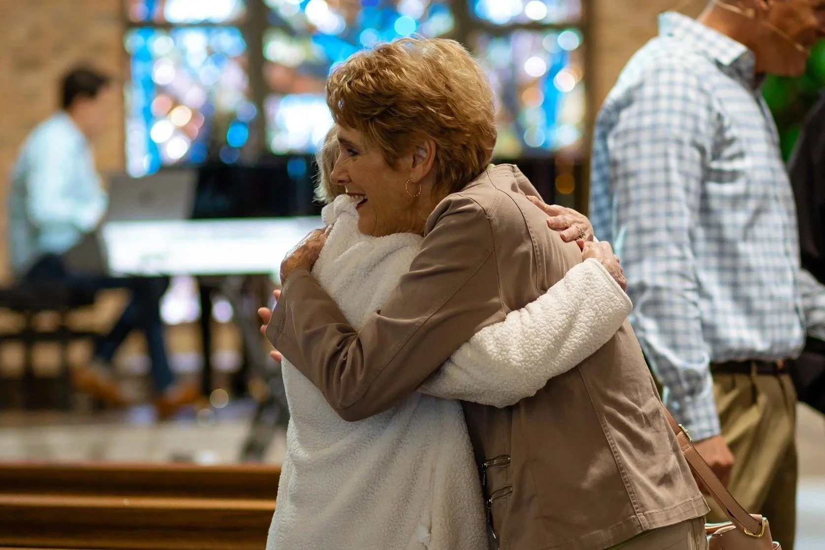 Two women hugging warmly inside a church or community hall, with blurred background of other people and stained glass windows.