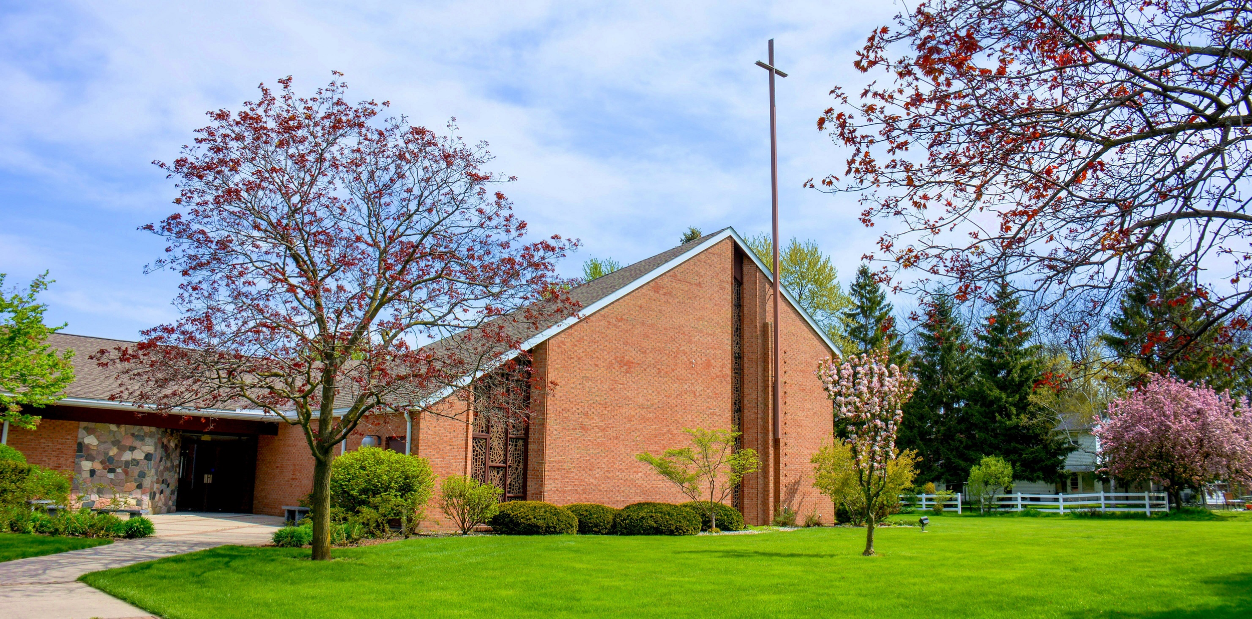 A church with a brick exterior and a tall steeple, surrounded by blooming trees and a well-maintained lawn under a partly cloudy sky.