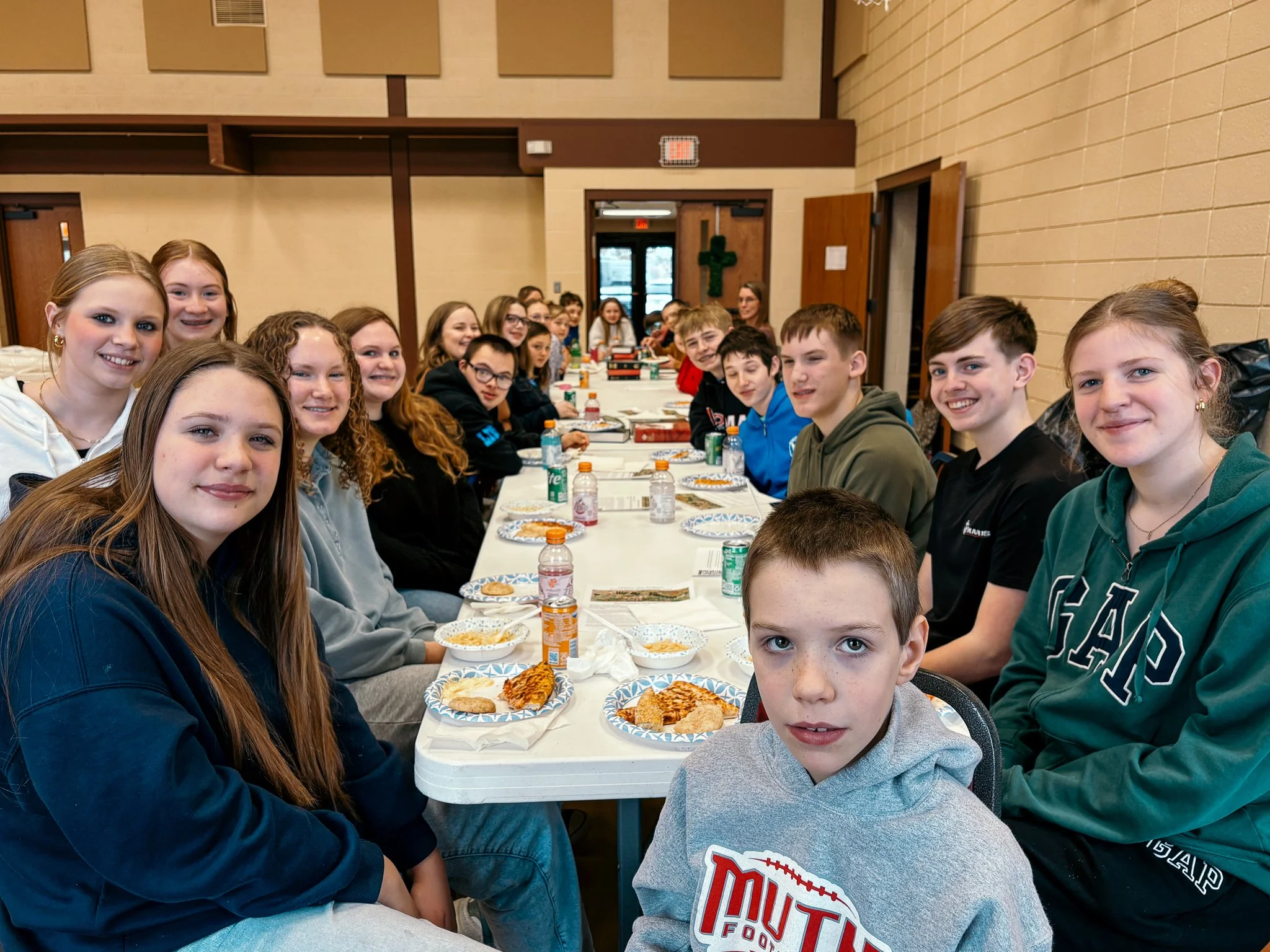 A group of children and teenagers sitting at a long table in a large room, enjoying a meal with slices of pizza and bottles of drinks.