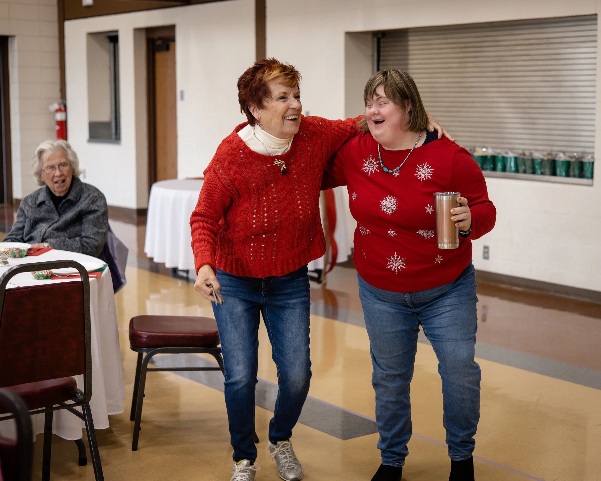 Two women laughing and embracing at a holiday gathering, wearing festive sweaters with snowflake and deer motifs, with elderly women seated at tables in the background.