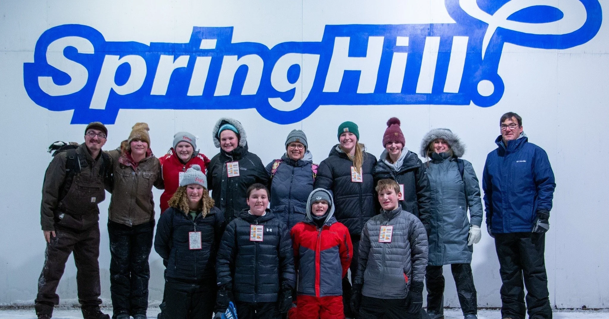 Group of children and adults in winter clothing standing in front of a large blue sign that says 'SpringHill' on a white wall.