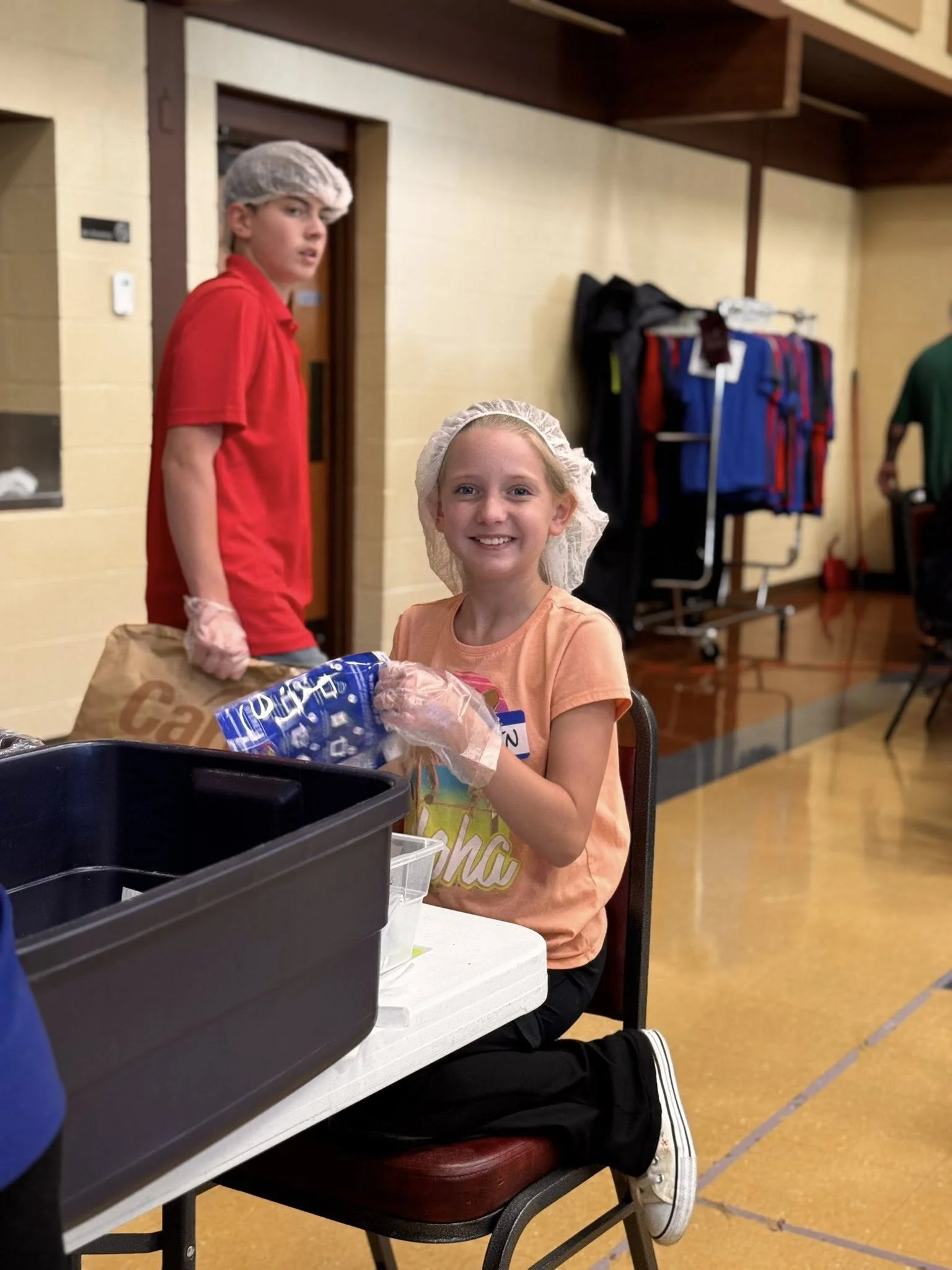 Two children wearing hairnets sitting at a table in a cafeteria or similar setting, with one smiling and the other looking to the side. A bag of chips and a black container are on the table. In the background, there are coat racks with clothing and a