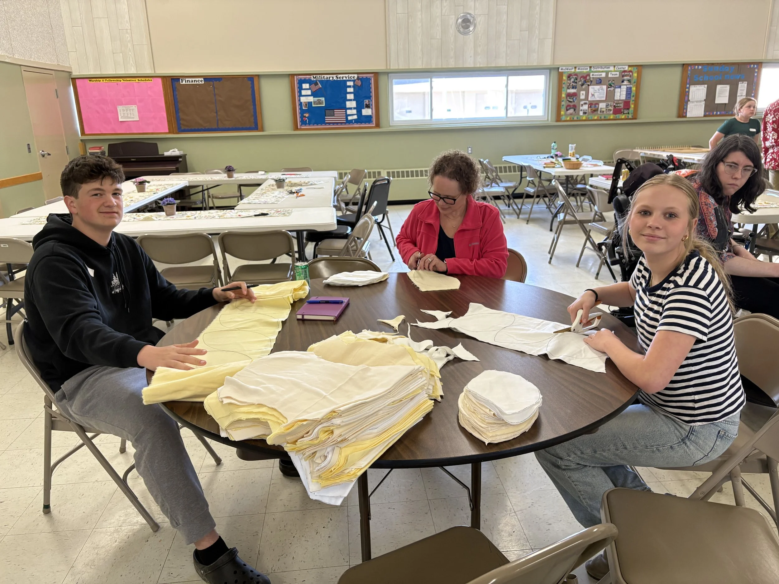 Four people sitting at a round table sorting yellow fabric or paper, with stacks of similar material in front of each person. The background shows a bulletin board with various posters and windows.