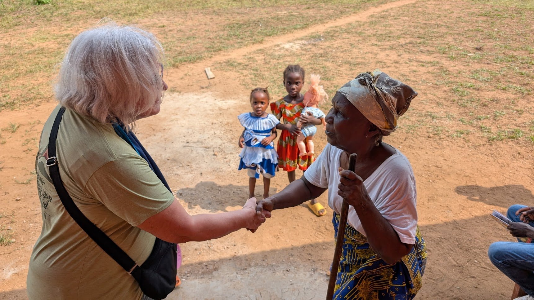 A woman with white hair shaking hands with an elderly woman wearing a headscarf, while three young girls stand behind them on a dirt ground amidst grass.