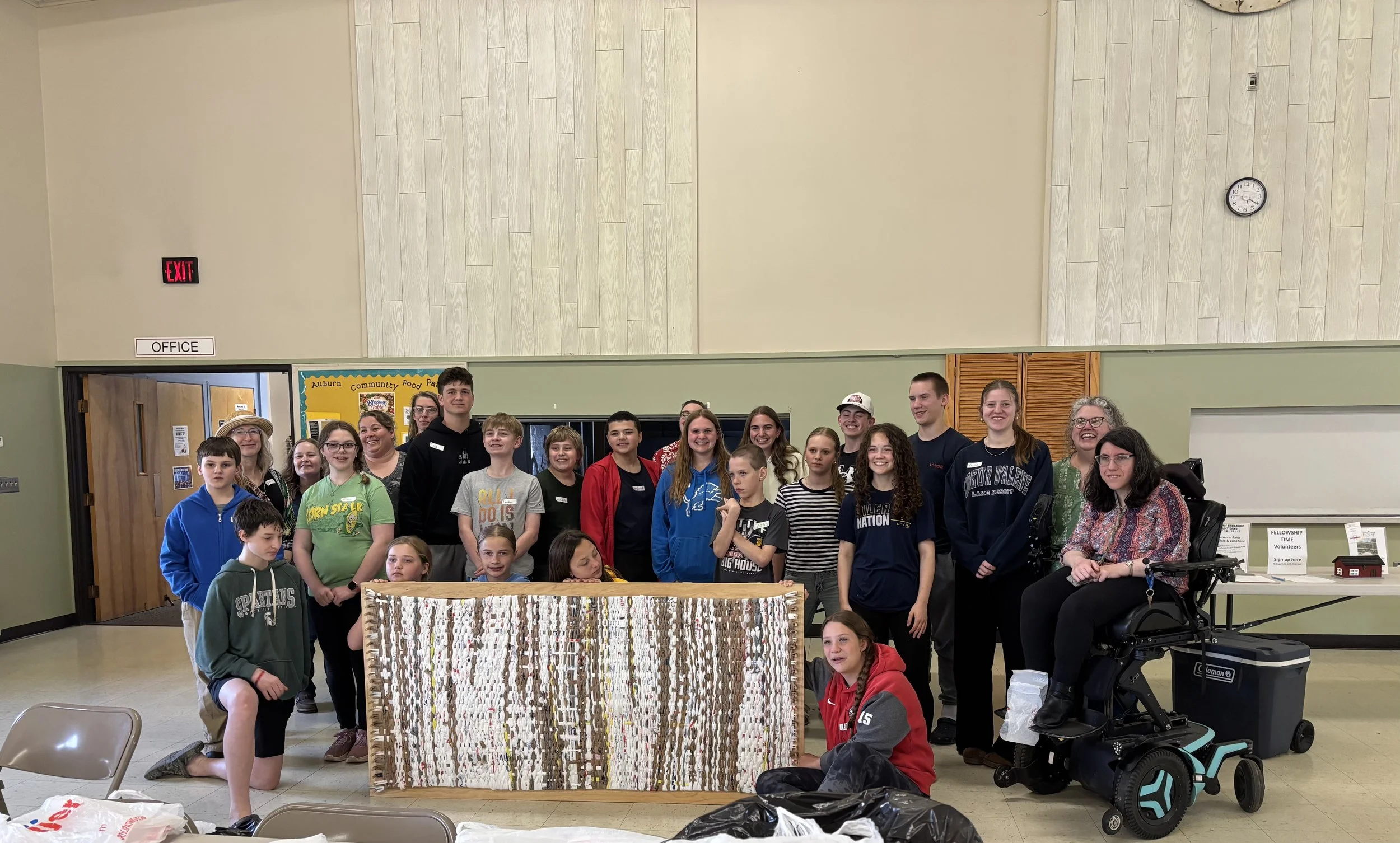 Group of children and adults posing inside a community center. The background includes a bulletin board, an exit sign, and a large clock. The group is gathered around a woven artwork displayed on a table.