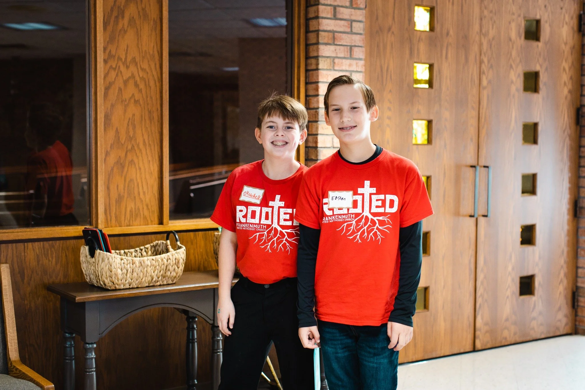 Two boys standing indoors in front of a wooden wall with small square windows, wearing matching red church t-shirts with white text and graphics, smiles on their faces.