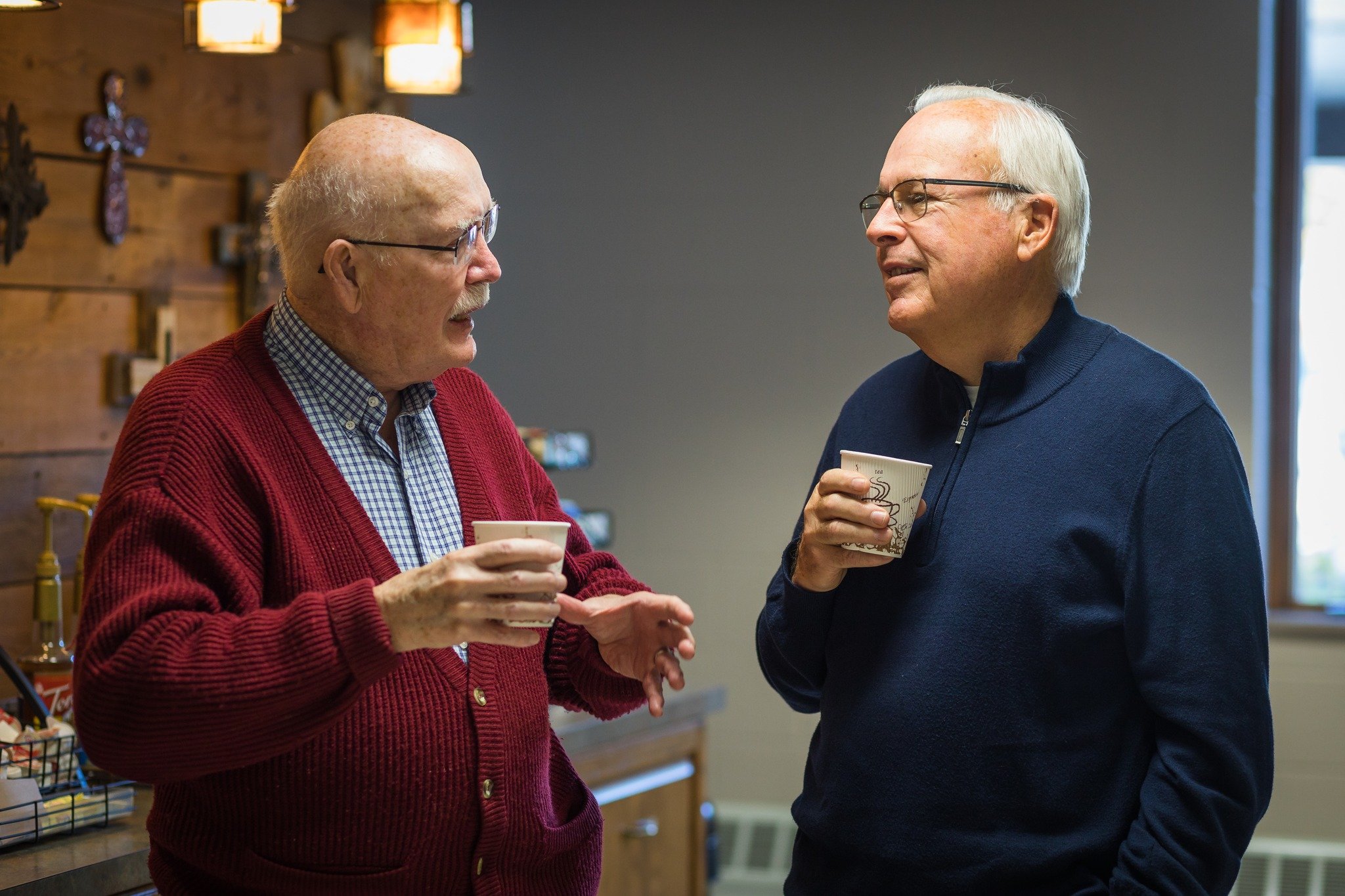 Two older men with glasses are talking and holding coffee cups while standing in church lounge.
