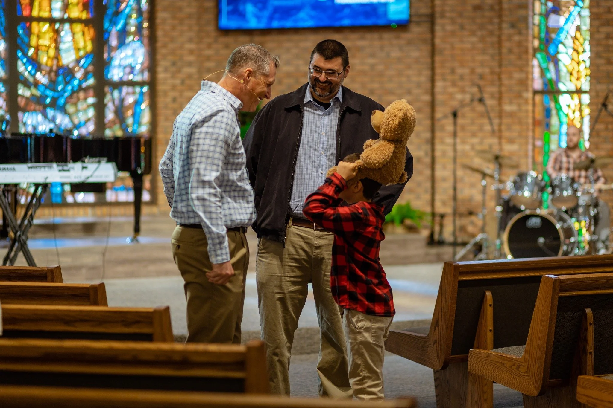 Three men and a boy with a teddy bear inside a church. The men appear to be having a pleasant conversation, smiling at each other. The background shows stained glass windows and musical instruments.