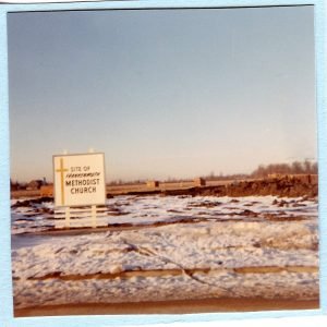 A sign on a snowy landscape indicating the site of a Methodist church, with an open field and a distant horizon under a clear sky.