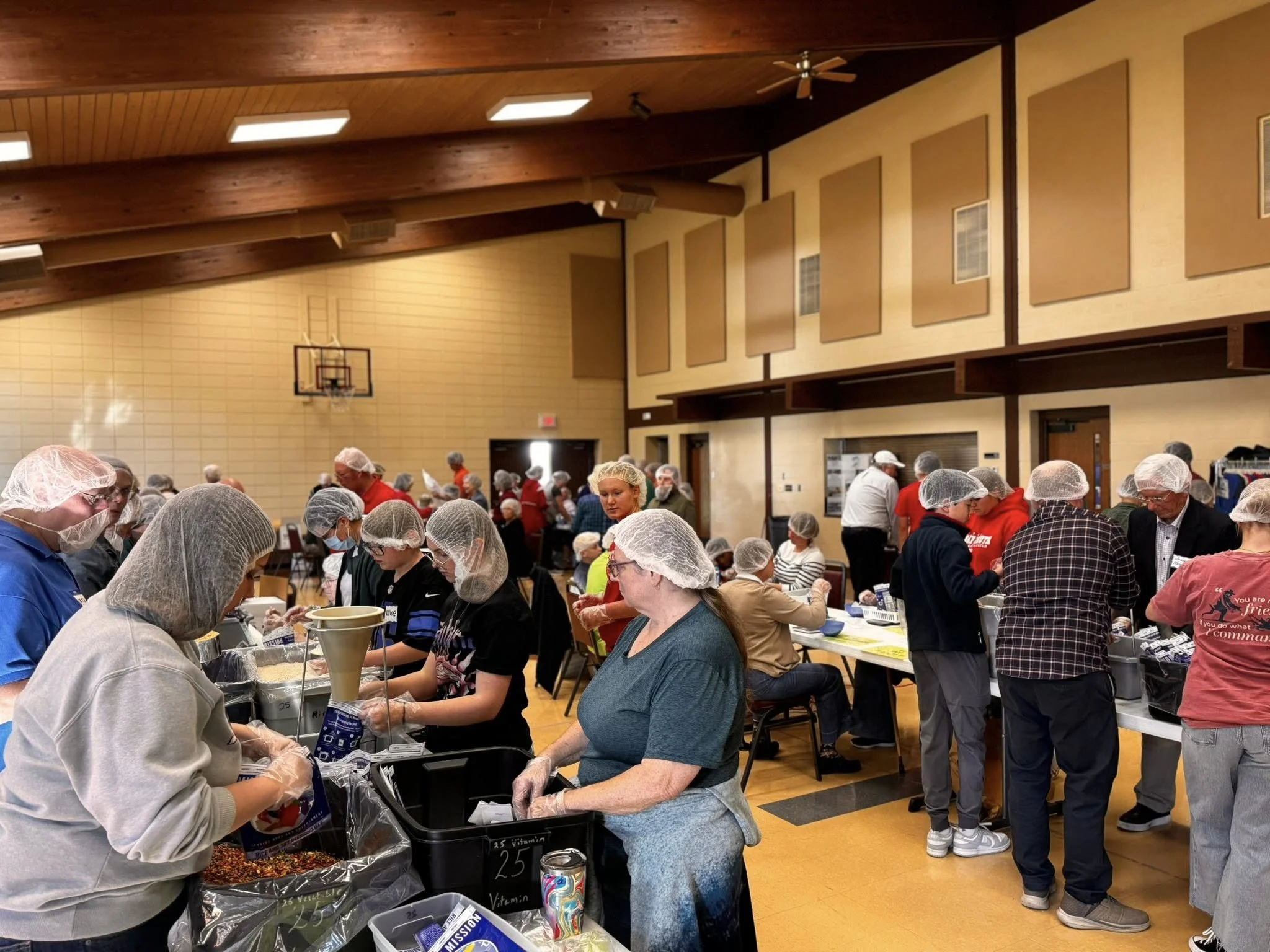 People wearing hairnets and masks gathered in a gymnasium-like space, packaging food for distribution near tables filled with bins of food items.