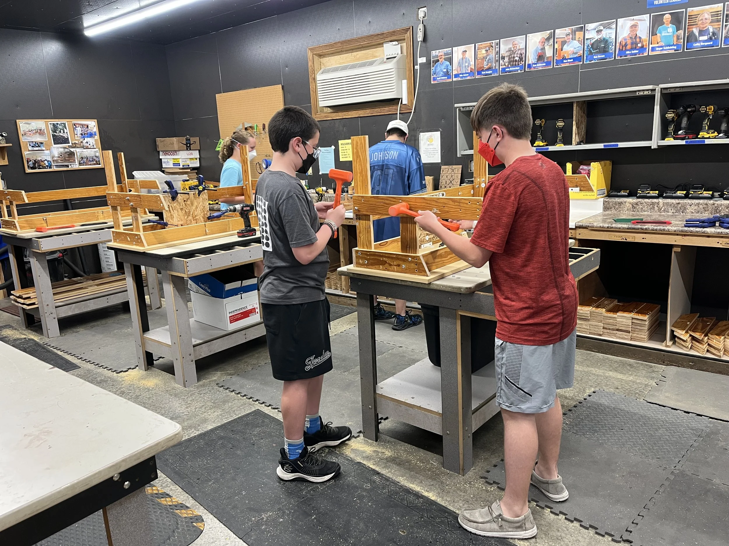 Two boys wearing face masks working on woodworking projects at a workshop table, with shelves of tools and photos on the walls behind them.