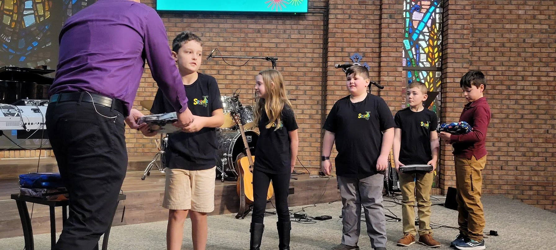 A man in a purple shirt is presenting awards or certificates to five children standing on a stage in front of a brick wall and stained glass windows. The children are dressed casually, some holding items, and are in line receiving recognition. Musica