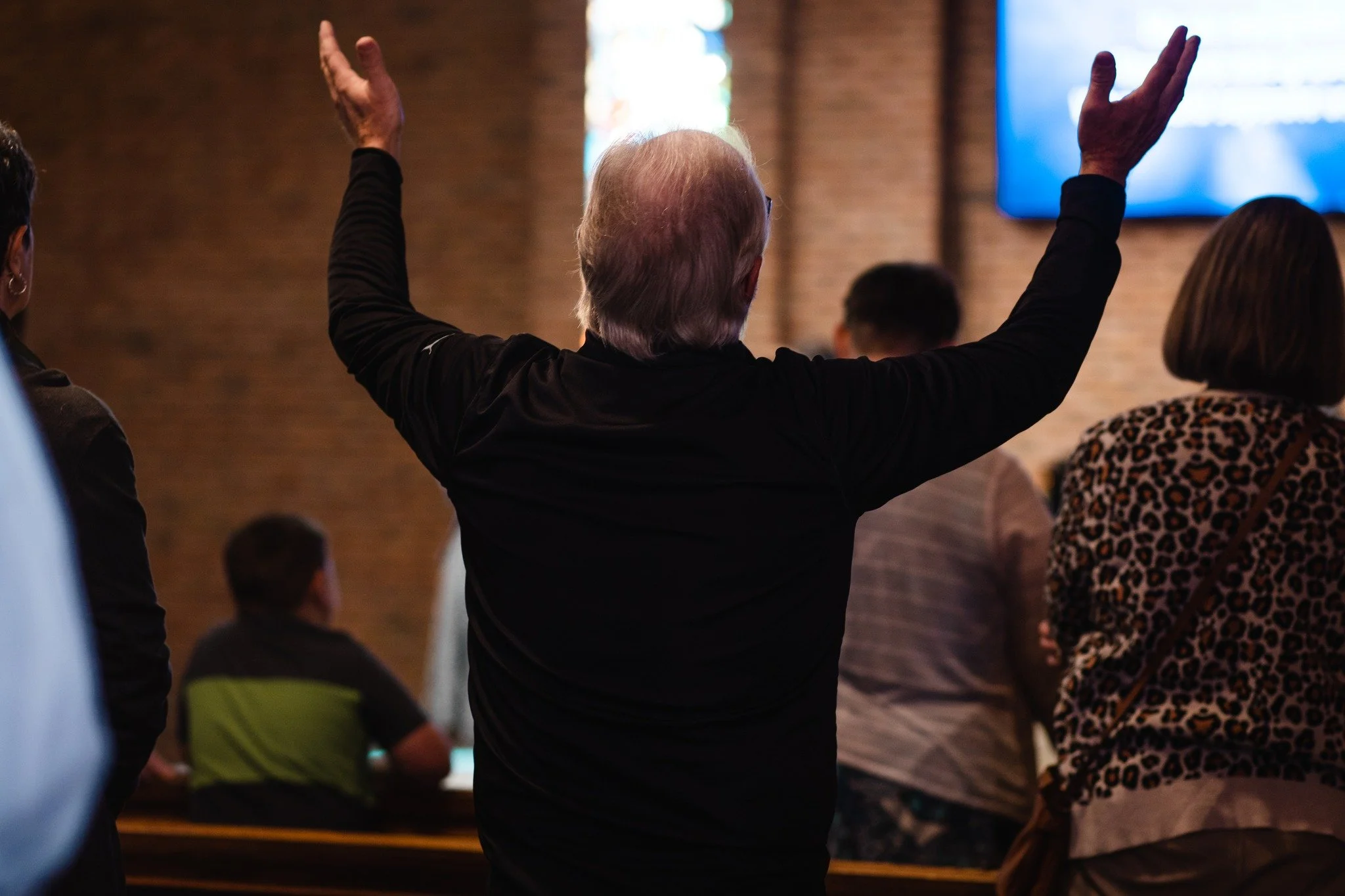 A person with gray hair and wearing glasses is raising both arms in a church or conference setting, with other people seated and a large screen visible in the background.