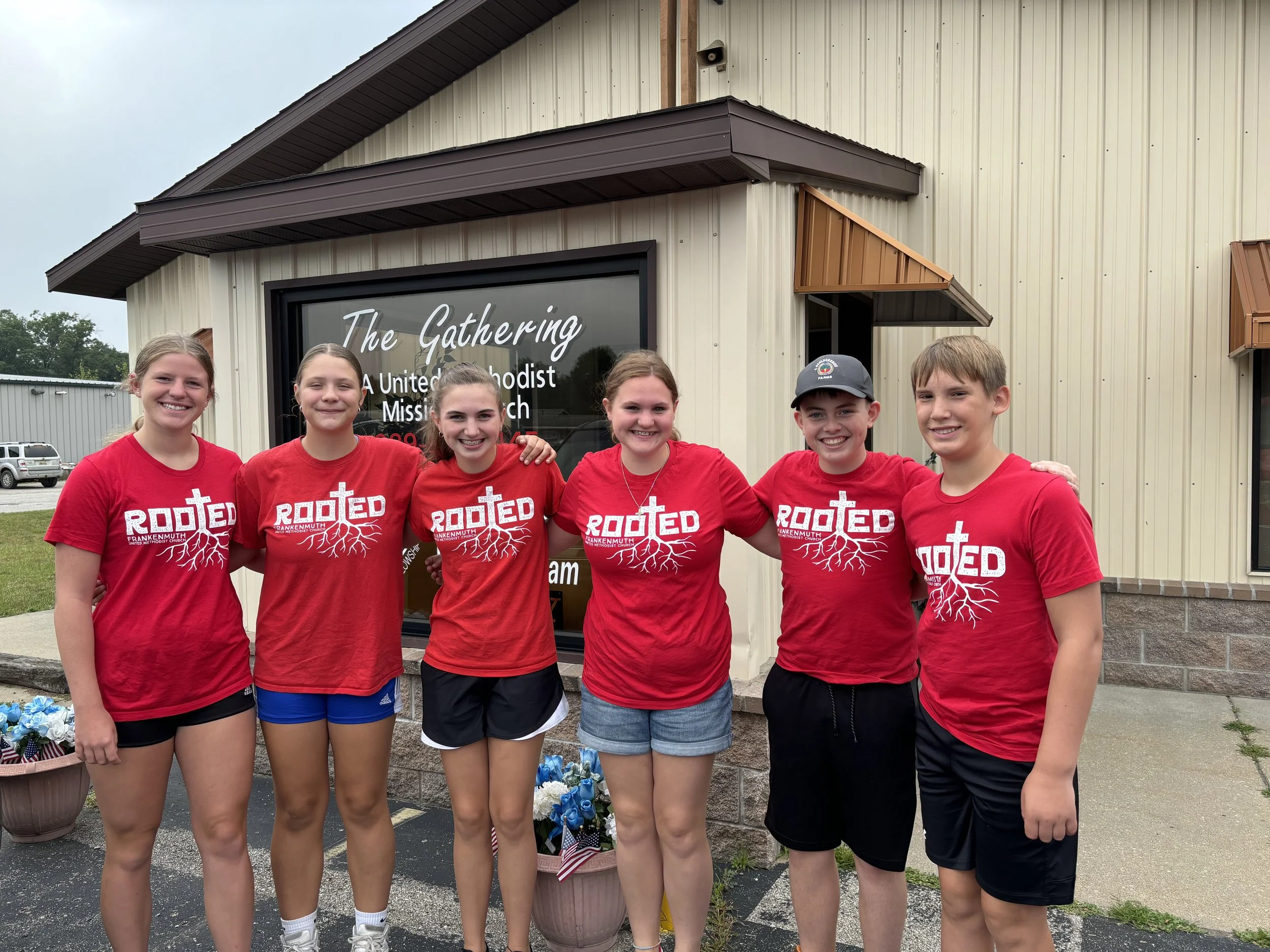 Six children wearing red shirts with the word 'Rooted' and a tree design stand outside a building with a sign that reads 'The Gathering United Methodist Church.' They are smiling and standing close together, with some placing their arms around each other.