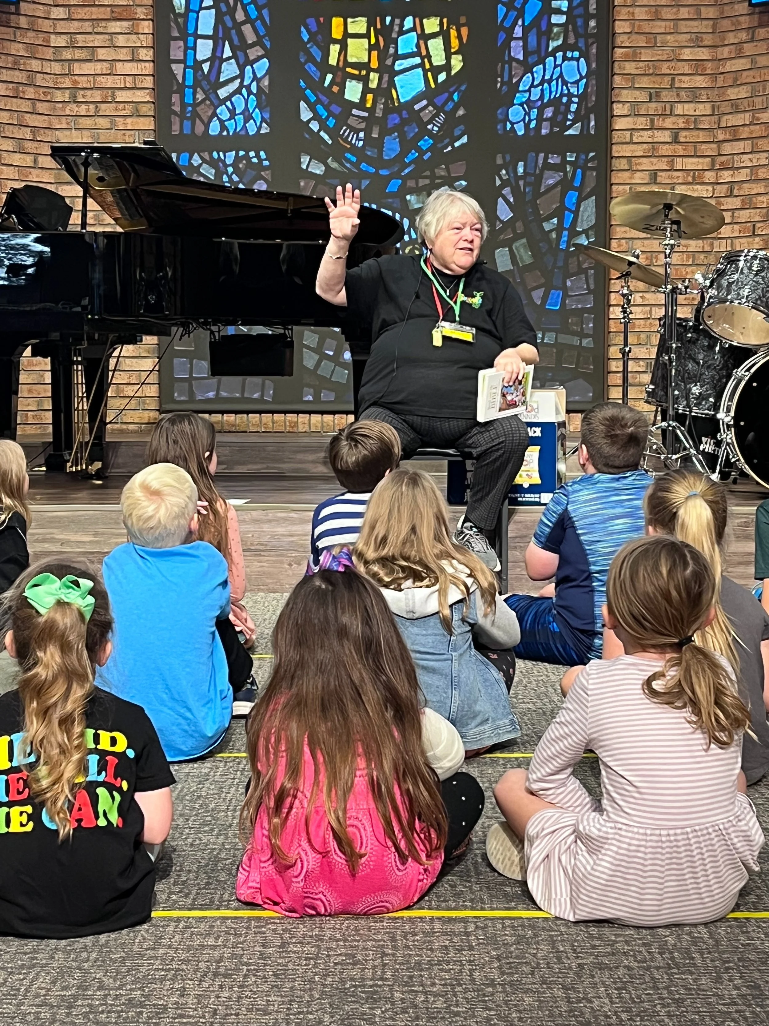 An older woman with short white hair, sitting in front of children and gesturing with her right hand during a presentation or reading session, with a stained-glass window in the background, a black piano on the left, and a drum set on the right in a 