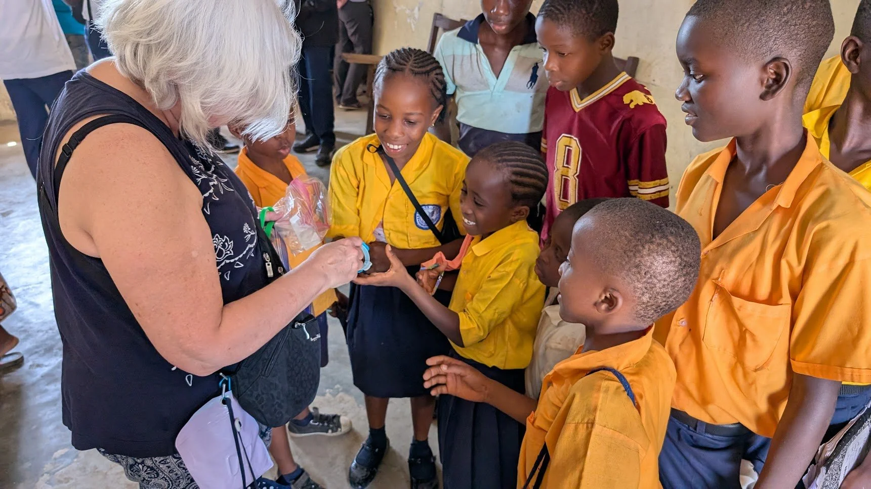 An older woman with gray hair giving a small gift to a group of school children dressed in yellow uniforms, inside a building with a concrete floor and walls. The children are smiling and gathered around her.