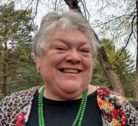A smiling elderly woman with short gray hair, wearing a black top, a green beaded necklace, and a floral shawl, outdoors near trees.