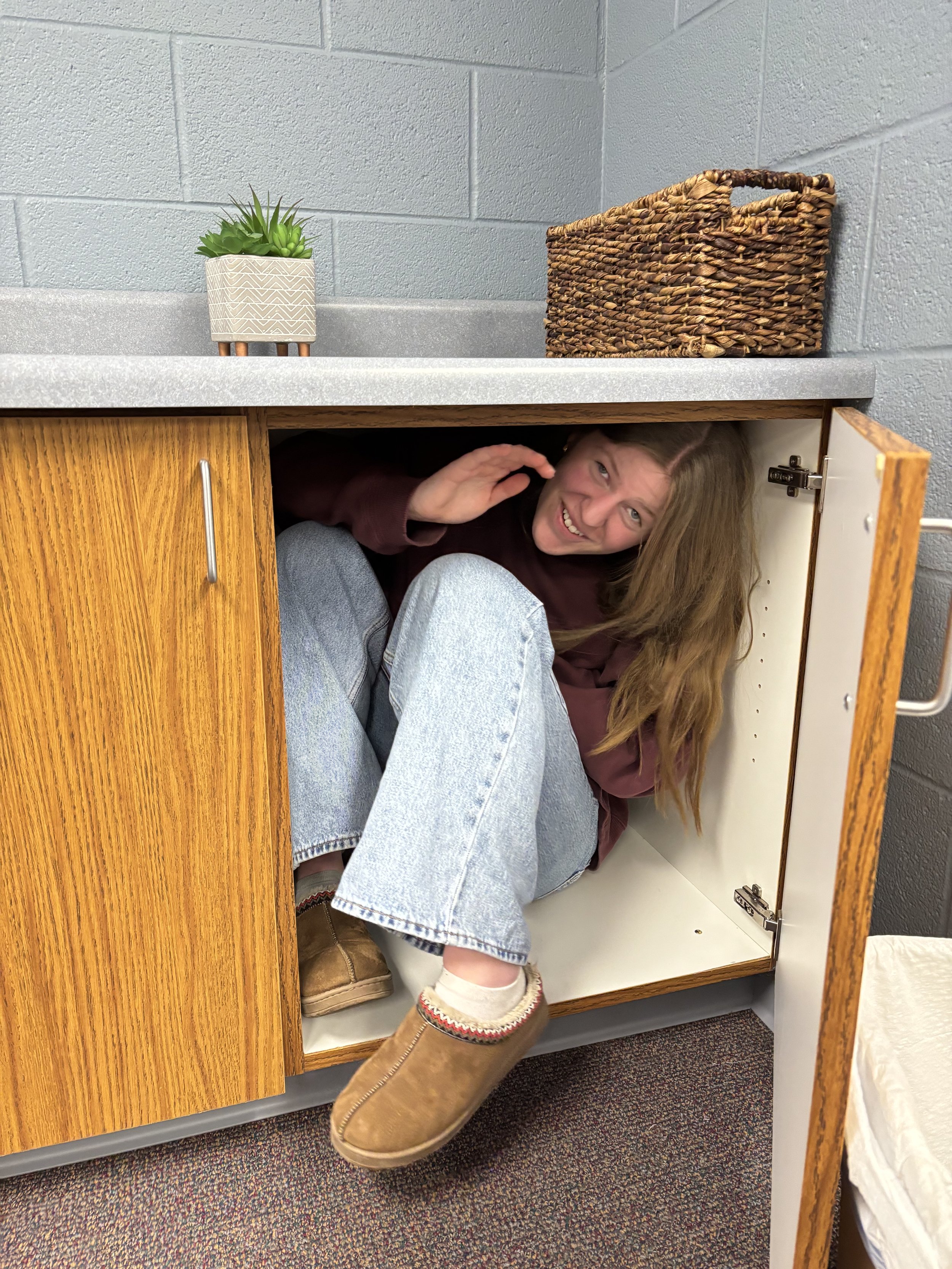 A woman crouching inside a cabinet with a surprised and playful expression, with her head and shoulders visible. The cabinet door is open, revealing her sitting inside.