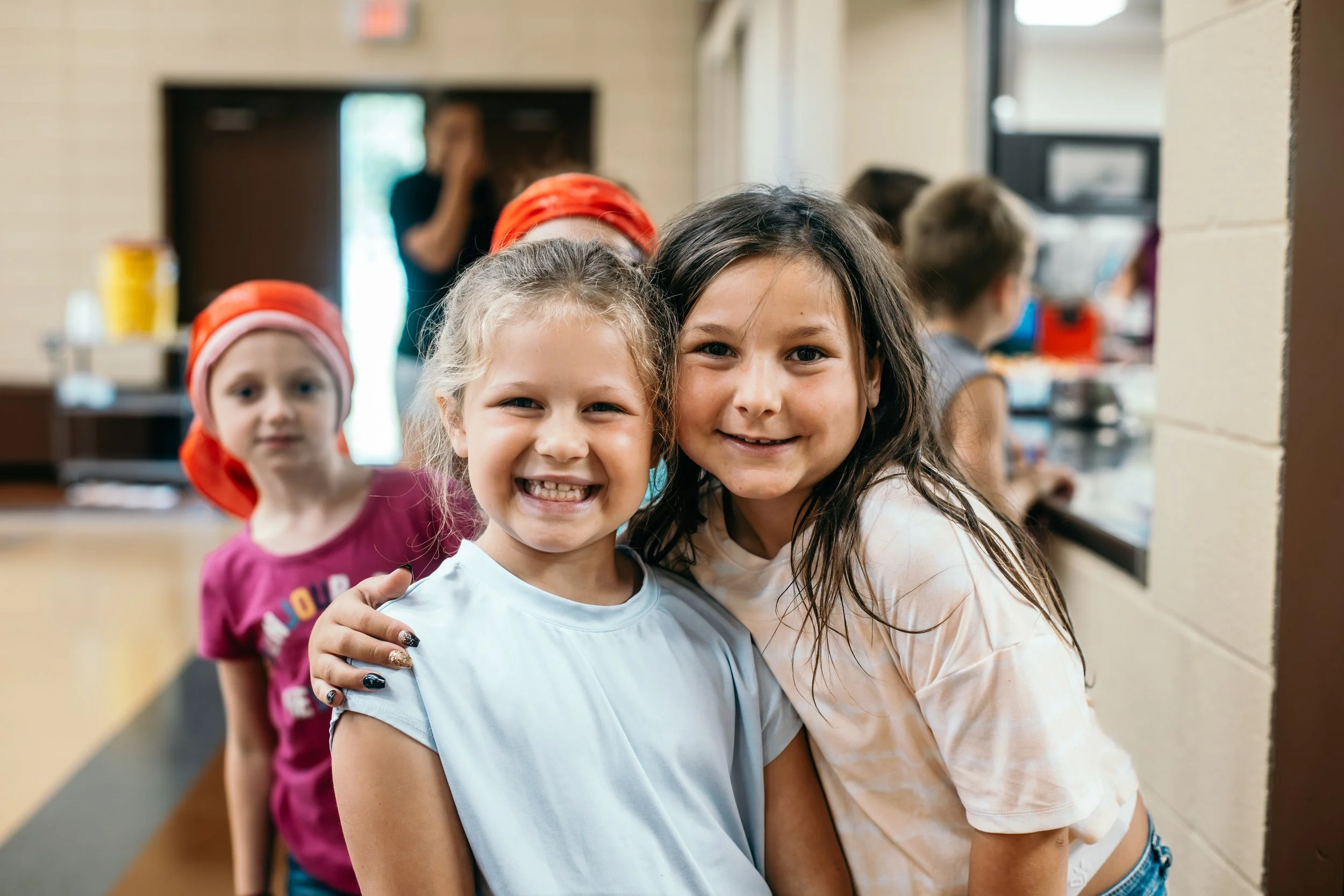 A group of young girls smiling and hugging in a line in what appears to be a community center or church hallway, with other children and a woman in the background.