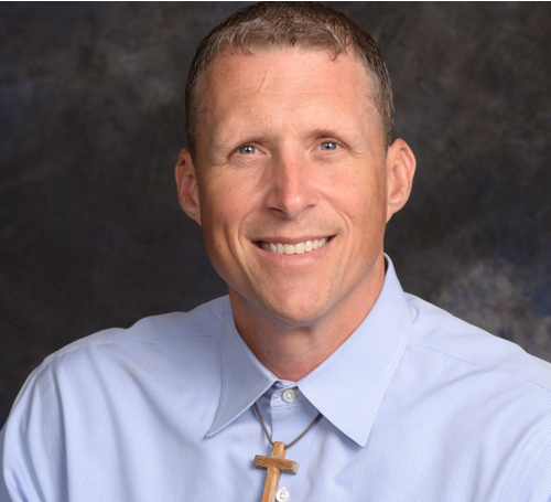 Smiling man wearing a light blue collared shirt and a wooden cross necklace, in front of a textured gray background.