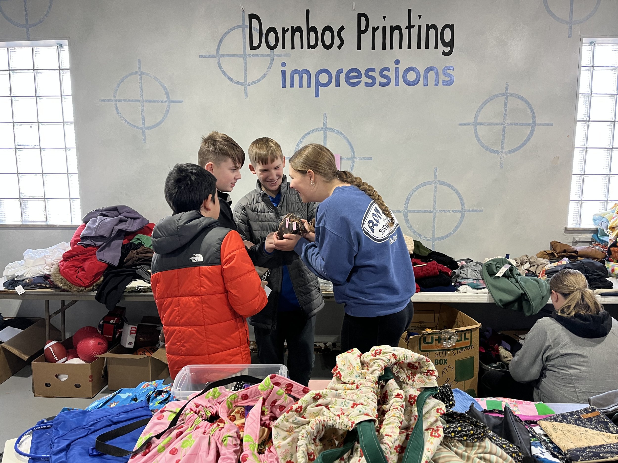 Group of people, including children and an adult, shopping for clothes and other items at a donation or thrift sale event. There are tables with various clothing, toys, and sports equipment.