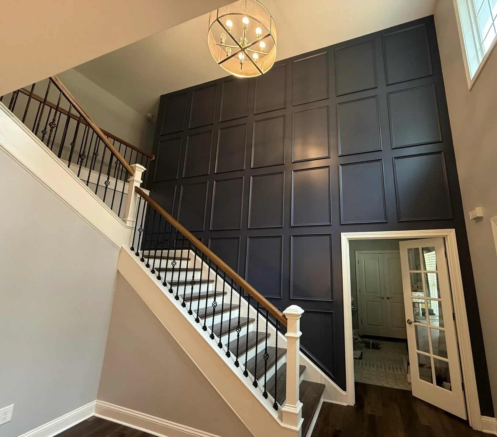 Interior view of a house with a staircase, dark paneled wall, chandelier, and an open doorway leading to another room.