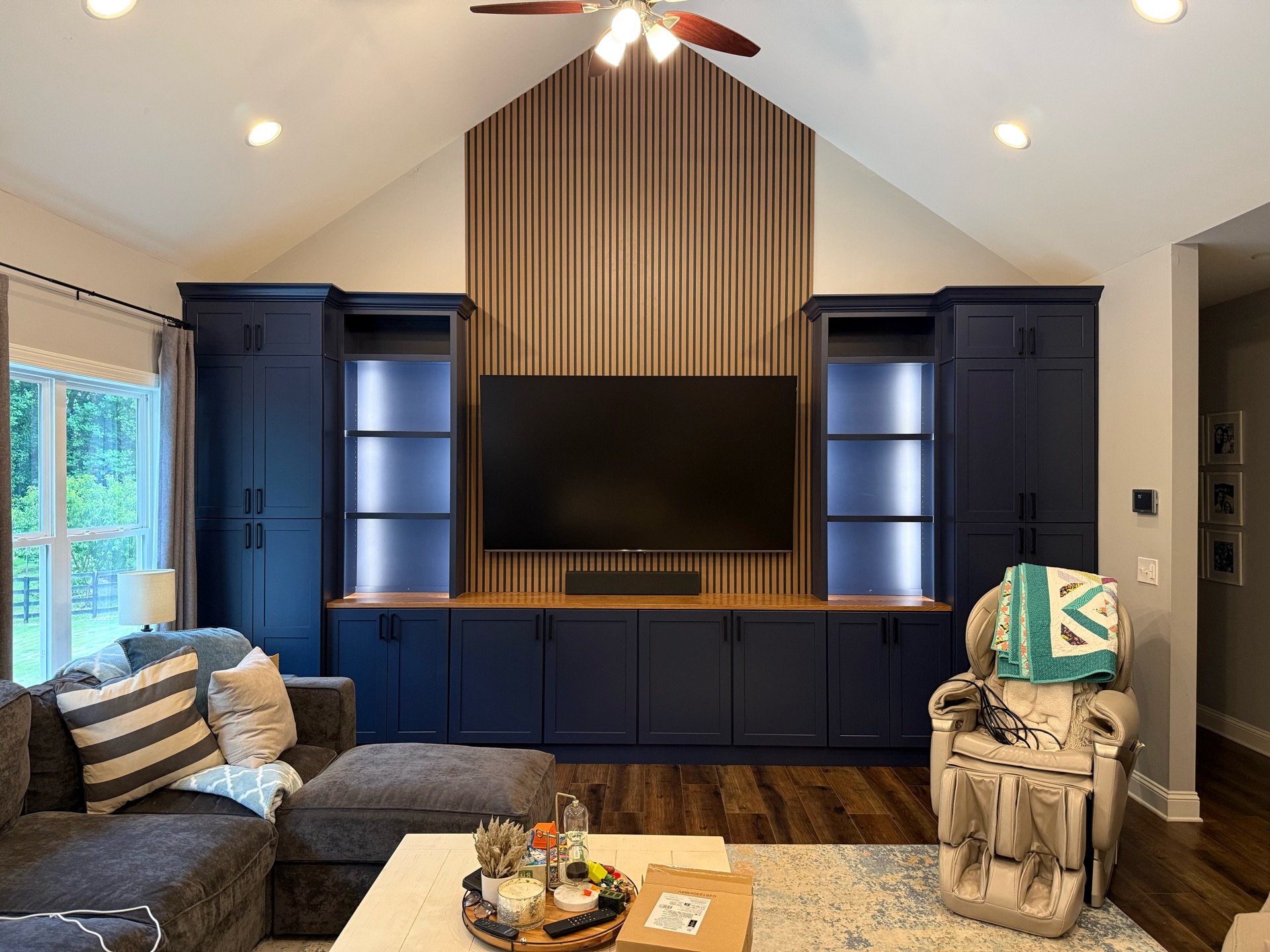 Living room with a dark blue built-in entertainment center featuring open shelves and closed cabinets, a large flat-screen TV mounted in the center, a window with curtains on the left, a beige chair with a colorful quilt draped over it on the right, 