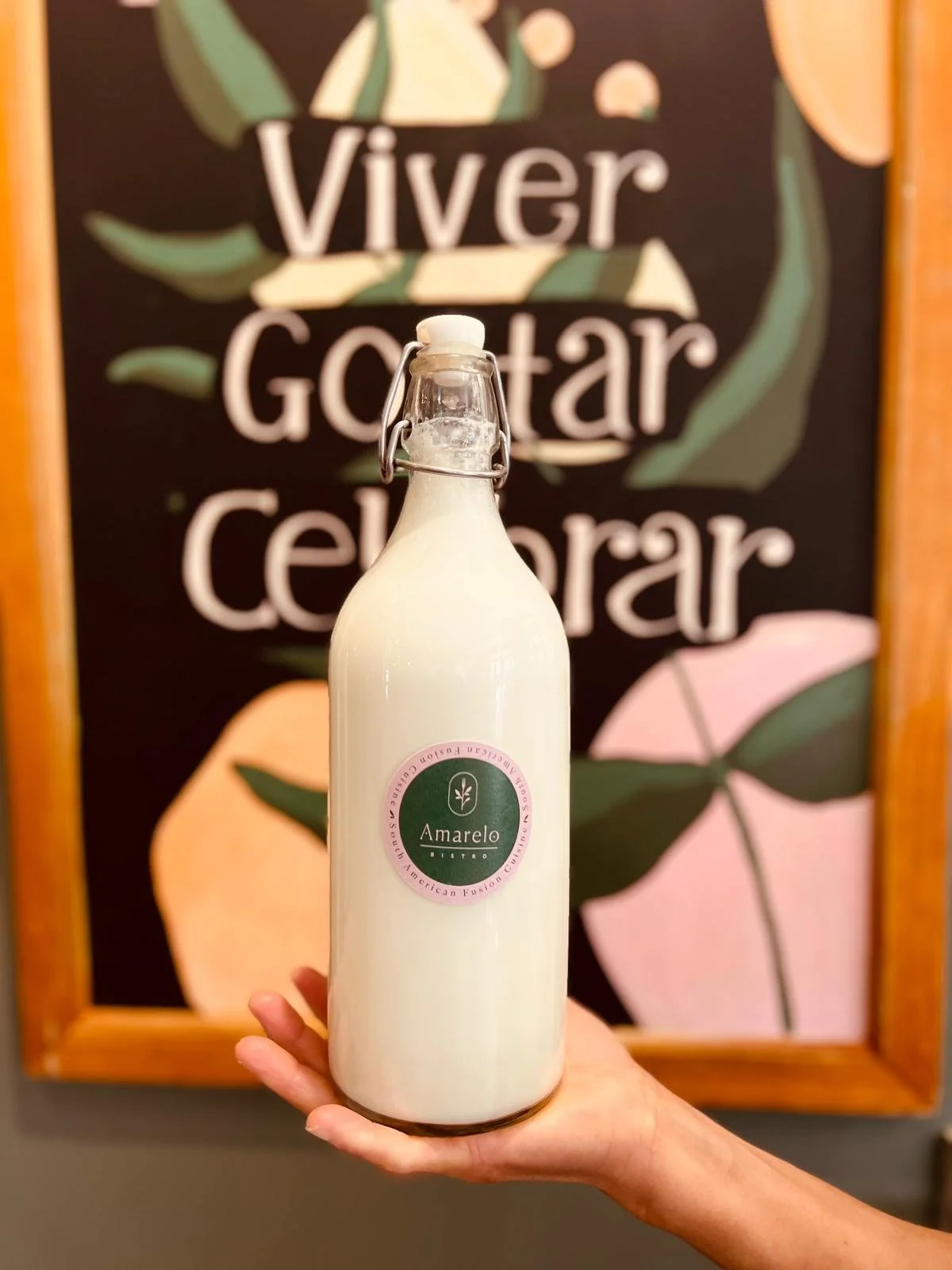 Person holding a bottle of Amarelo branded beverage in front of a black chalkboard with handwritten Spanish phrases about living, eating, and celebrating.