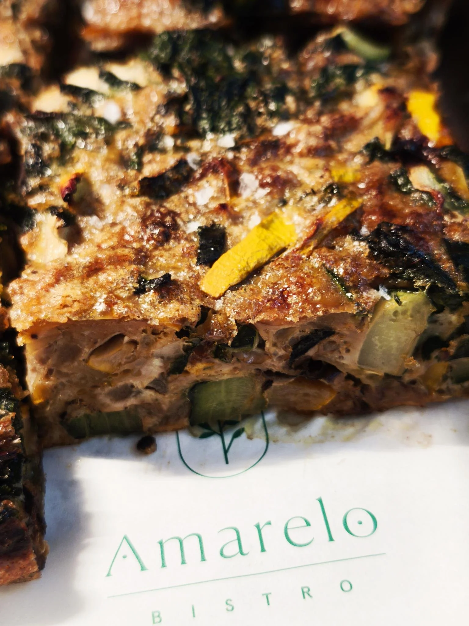 Close-up of a slice of vegetable frittata with zucchini, yellow peppers, and spinach on 'Amarelo Bistro' branded grease proof paper.