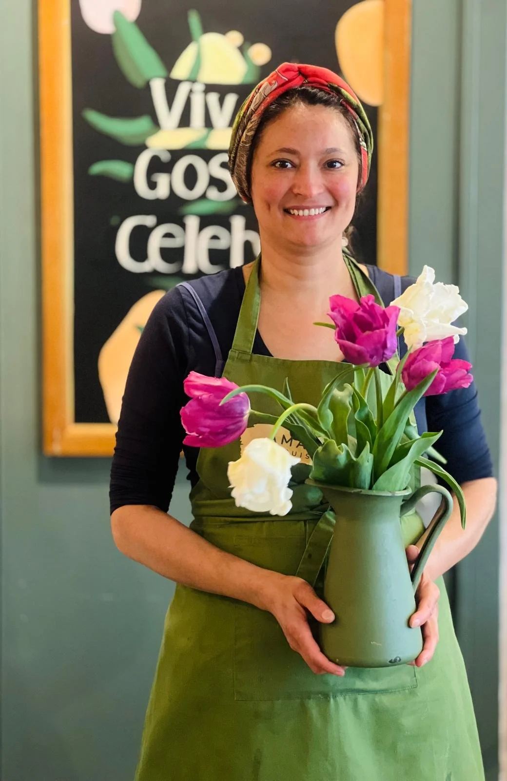 A woman standing indoors, smiling, holding a green pitcher with pink and white tulips. She is wearing a dark top, a green apron, and a colorful headscarf. In the background, there is a sign with some text and illustrations.
