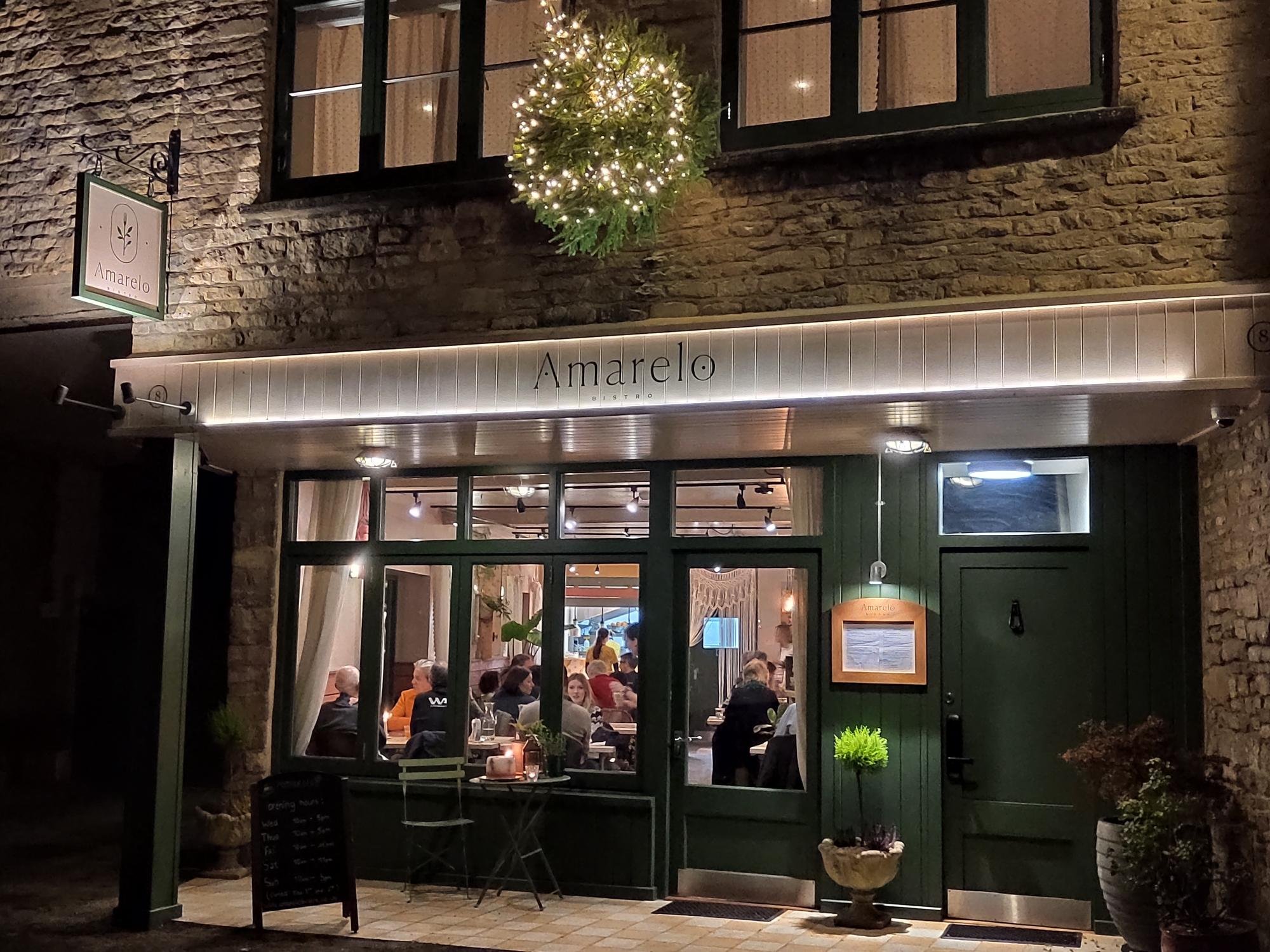 Christmas at Amarelo Bistro. An  exterior view of the restaurant at night, with a Cotswold stone facade, green door and window frames, a hanging wreath with lights, and people dining inside visible through the large windows.