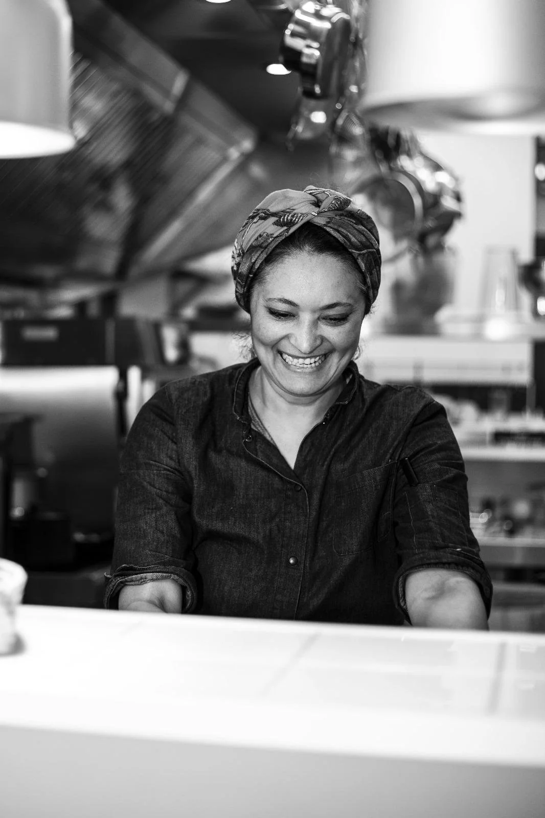 Black and white photo of a woman with a headscarf smiling in a kitchen or restaurant setting.