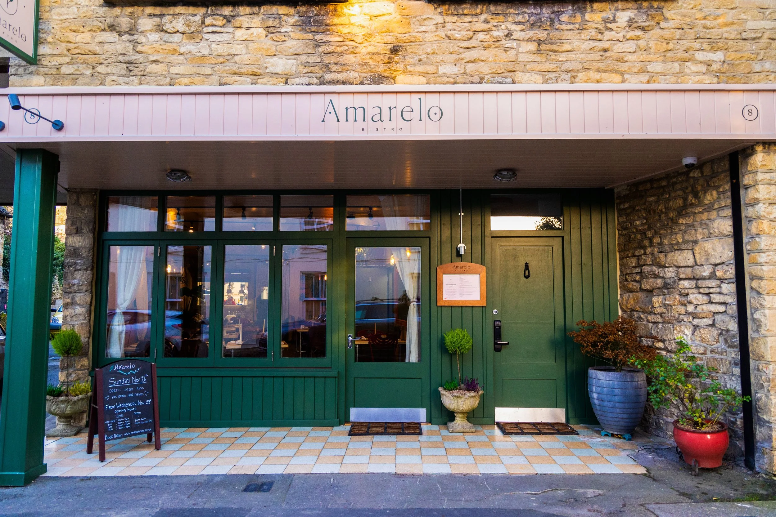 Front exterior of Amarelo Bistro with green entrance door, large windows, potted plants, chalkboard menu, and a pink awning