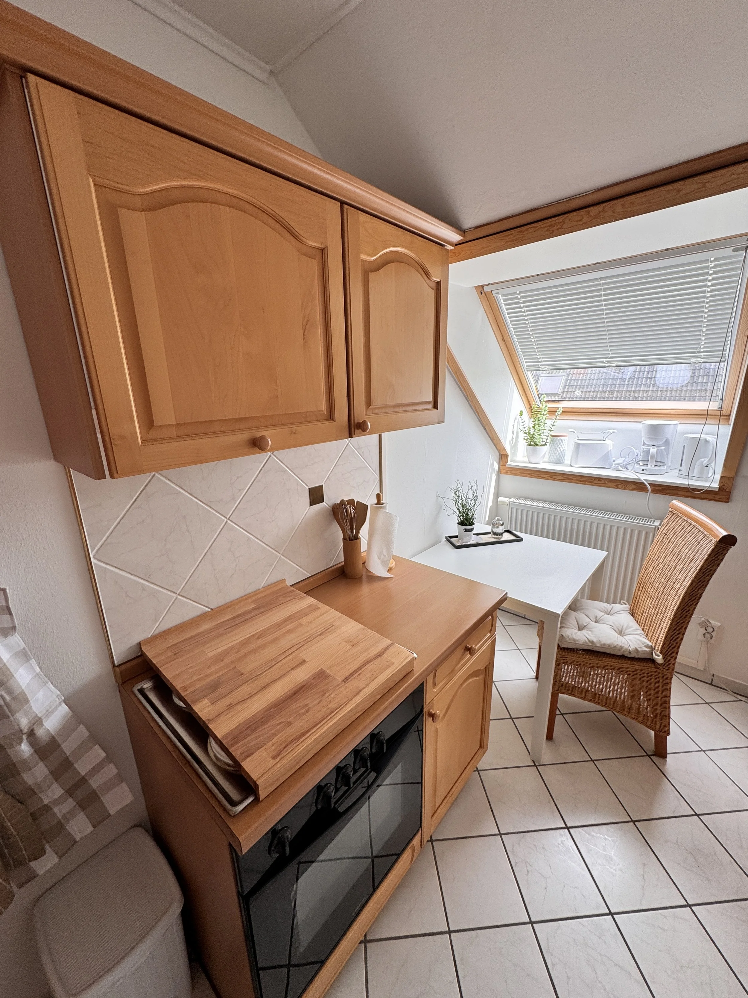 Kleine Küche mit Holzelementen, weißem Tisch, Stuhl mit Kissen, Fenster mit Rolladen, Pflanzen und Küchenutensilien.