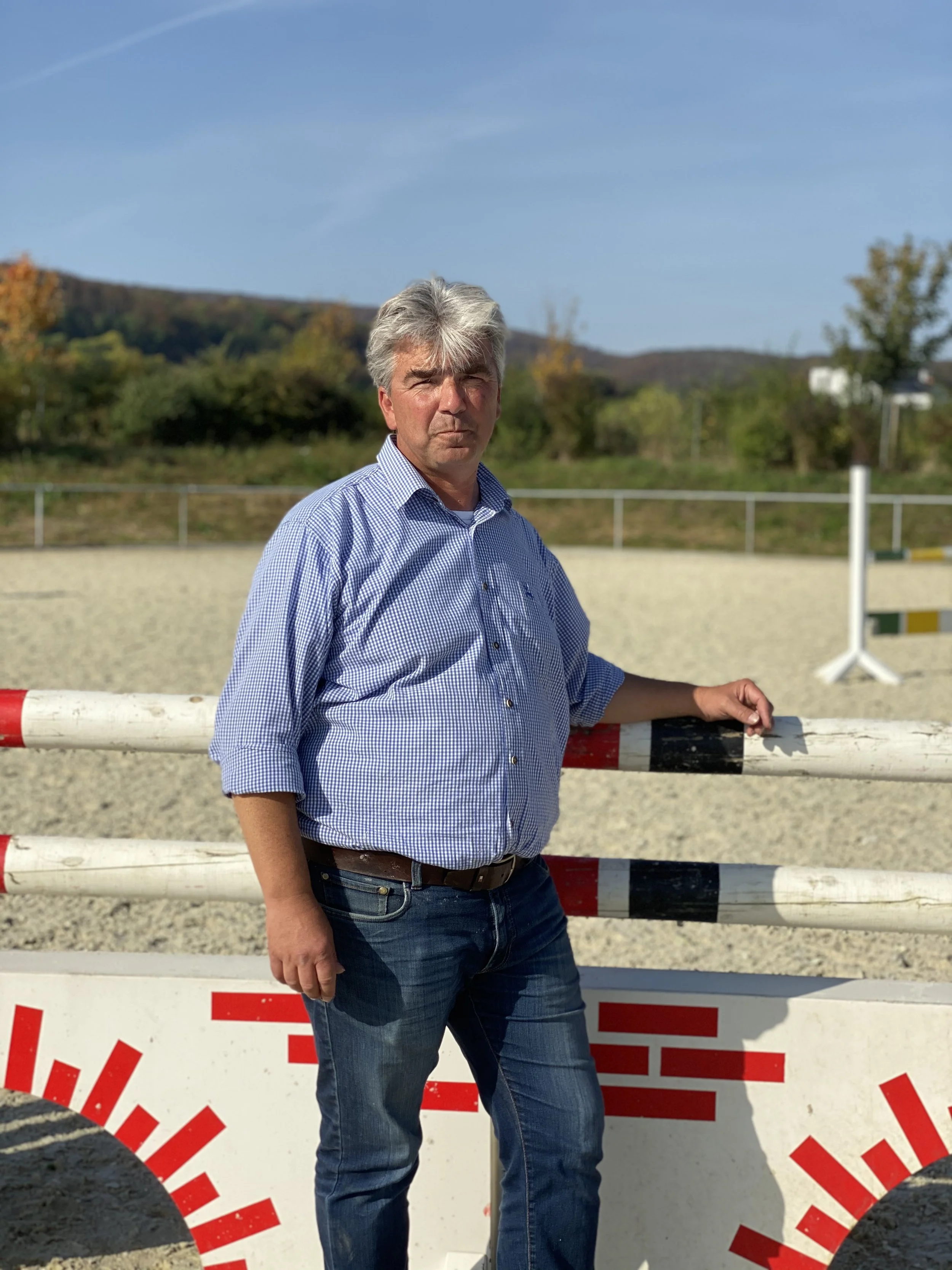 Mann in blauer kariert Hemd und Jeans lehnt an einer weißen Barriere auf einem Reitplatz bei sonnigem Wetter, im Hintergrund Bäume und Berge.
