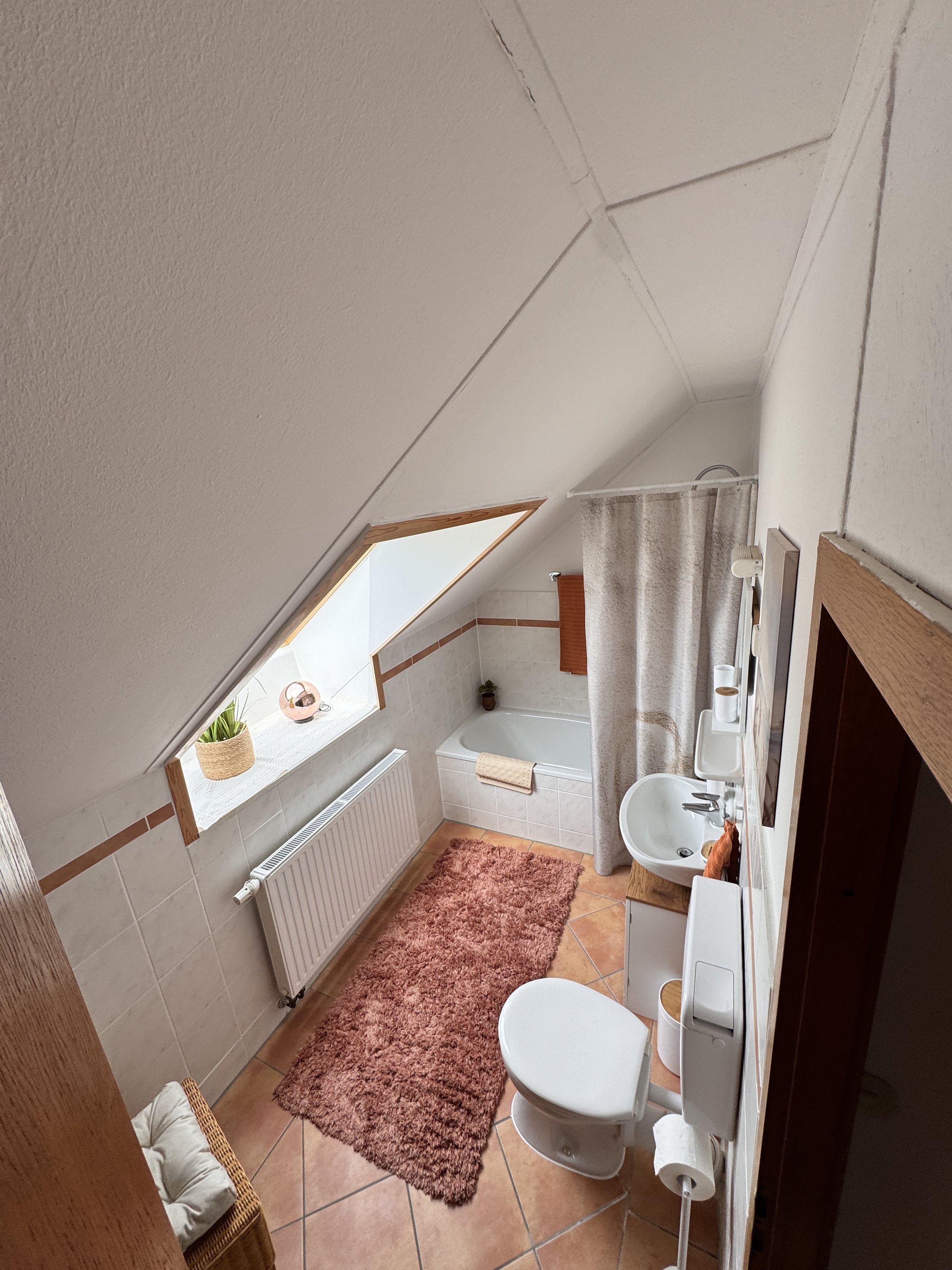 Ein kleines Badezimmer mit einer Toilette, einem Waschbecken, einer Badewanne und einem Fenster mit Vorhang. Die Wände sind gefliest, der Boden ist mit Terrakottafliesen ausgelegt. Es gibt einen braunen Teppich und einige Dekorationsartikel wie Pflan