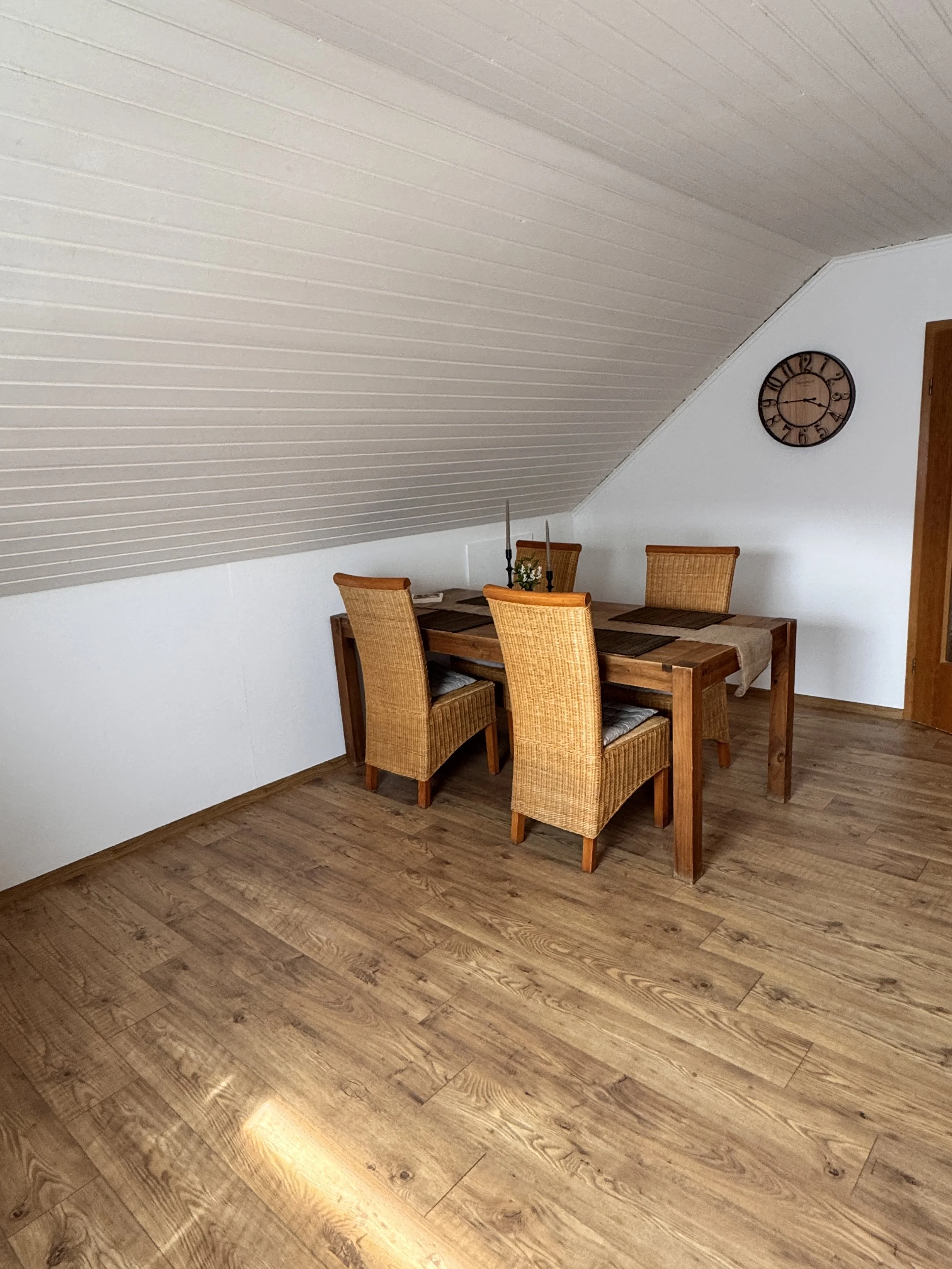 Esszimmer mit Holztisch, vier Stühlen, einer Tischdekoration mit Kerzen und einer Wanduhr an der weißen Wand, schräges Deckenpaneel, Holzboden.