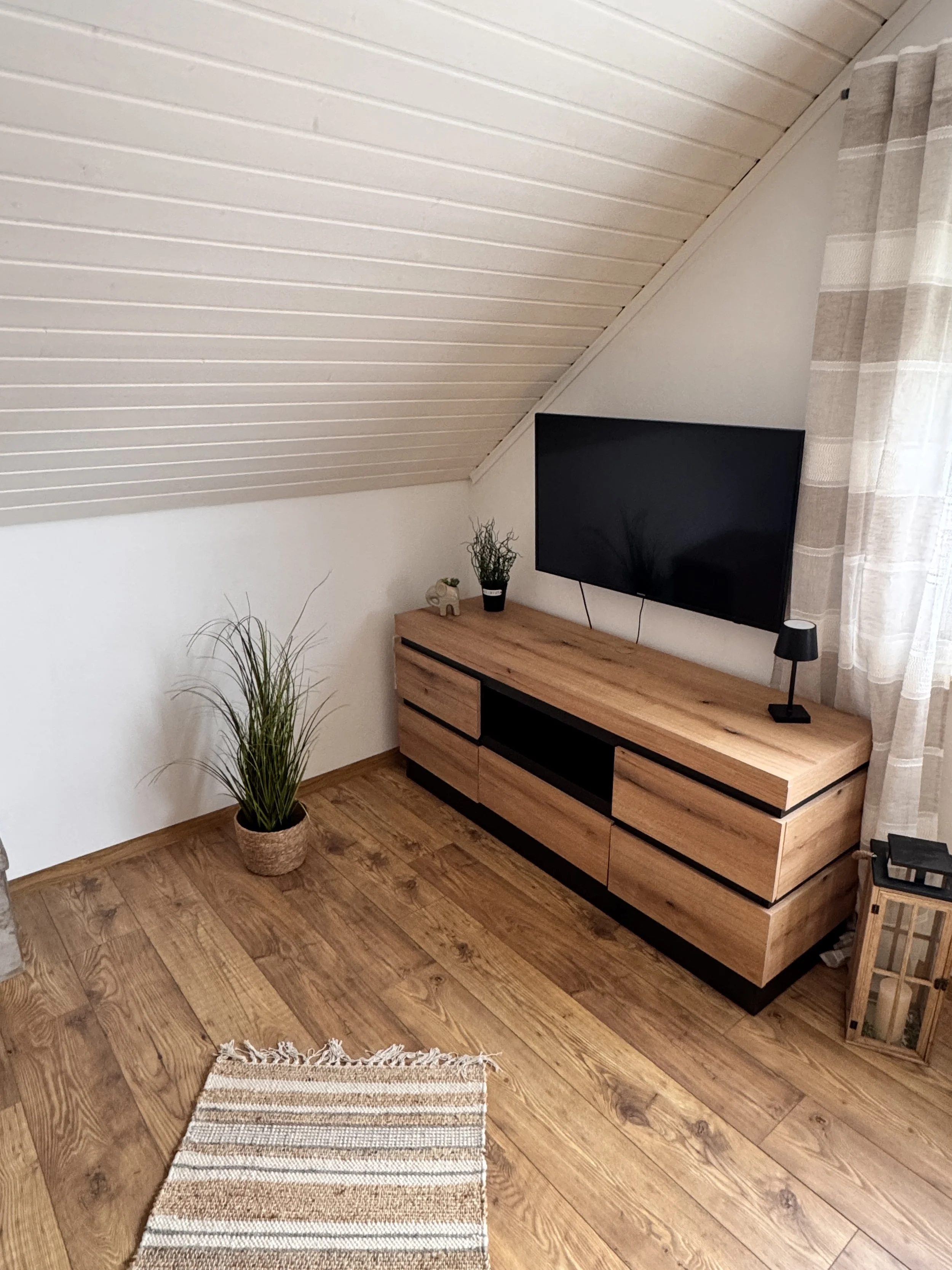 Ein minimalistisches Wohnzimmer mit einem Holz-TV-Schrank, einem Flachbildfernseher, mehreren Pflanzen, einem kleinen Tisch, einem Teppich und Gardinen vor einem Fenster.