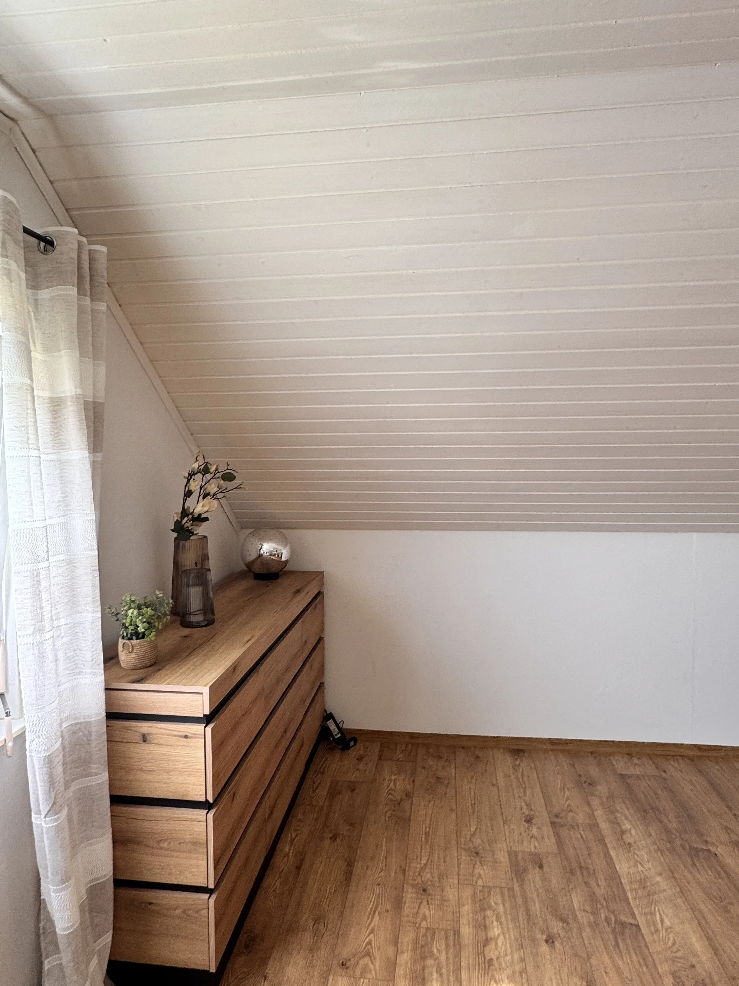 Ein Zimmer mit Holzfußboden, eine Kommode aus Holz mit drei Schubladen, dekoriert mit einer Vase mit Kunstblumen, einer kleinen Pflanze und einem dekorativen Gegenstand, daneben ein Fenster mit einem weißen Vorhang.