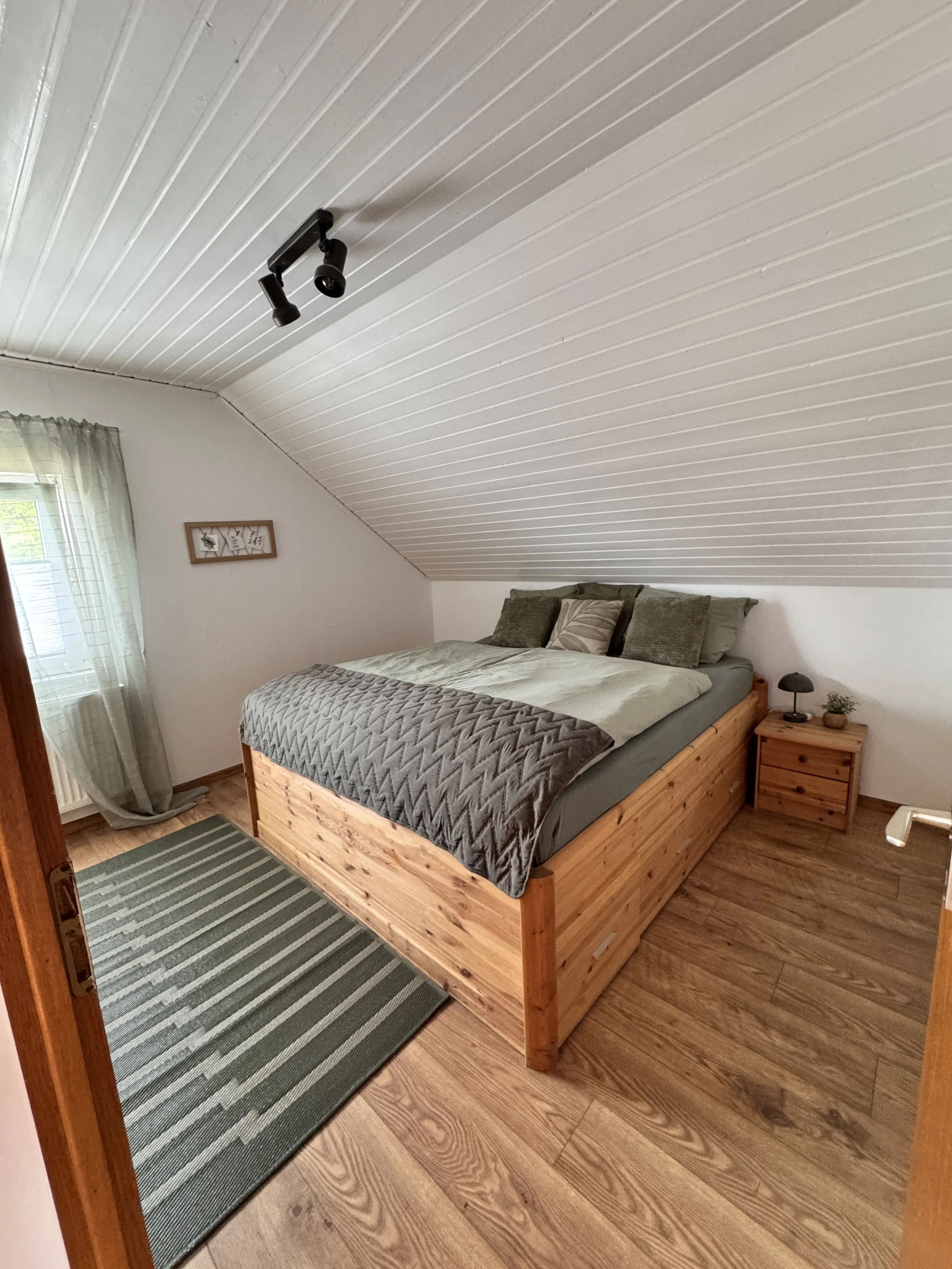 Schlafzimmer mit Holzbett, grau und beigem Bettzeug, seitlicher Nachttisch mit lampe und Pflanze, Holzboden, weiße Wand, schräg gespannte Decke mit drei Spotlights, Fenster mit grüner Gardine.