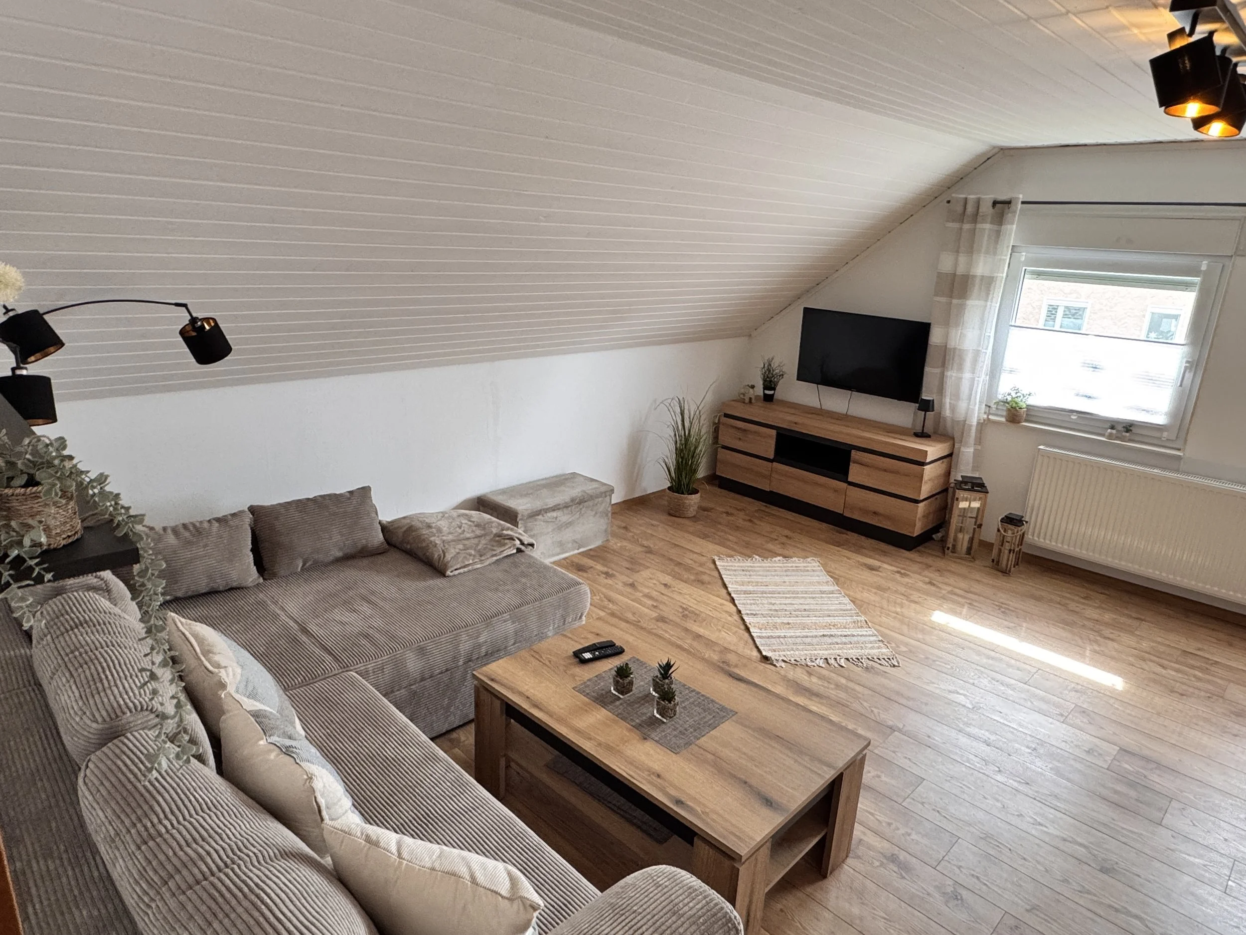 Gemütliches Wohnzimmer mit Couch, Couchtisch, Fernseher auf Holz-TV-Möbel, Fenster mit Vorhängen, Pflanzen und dekorative Gegenstände