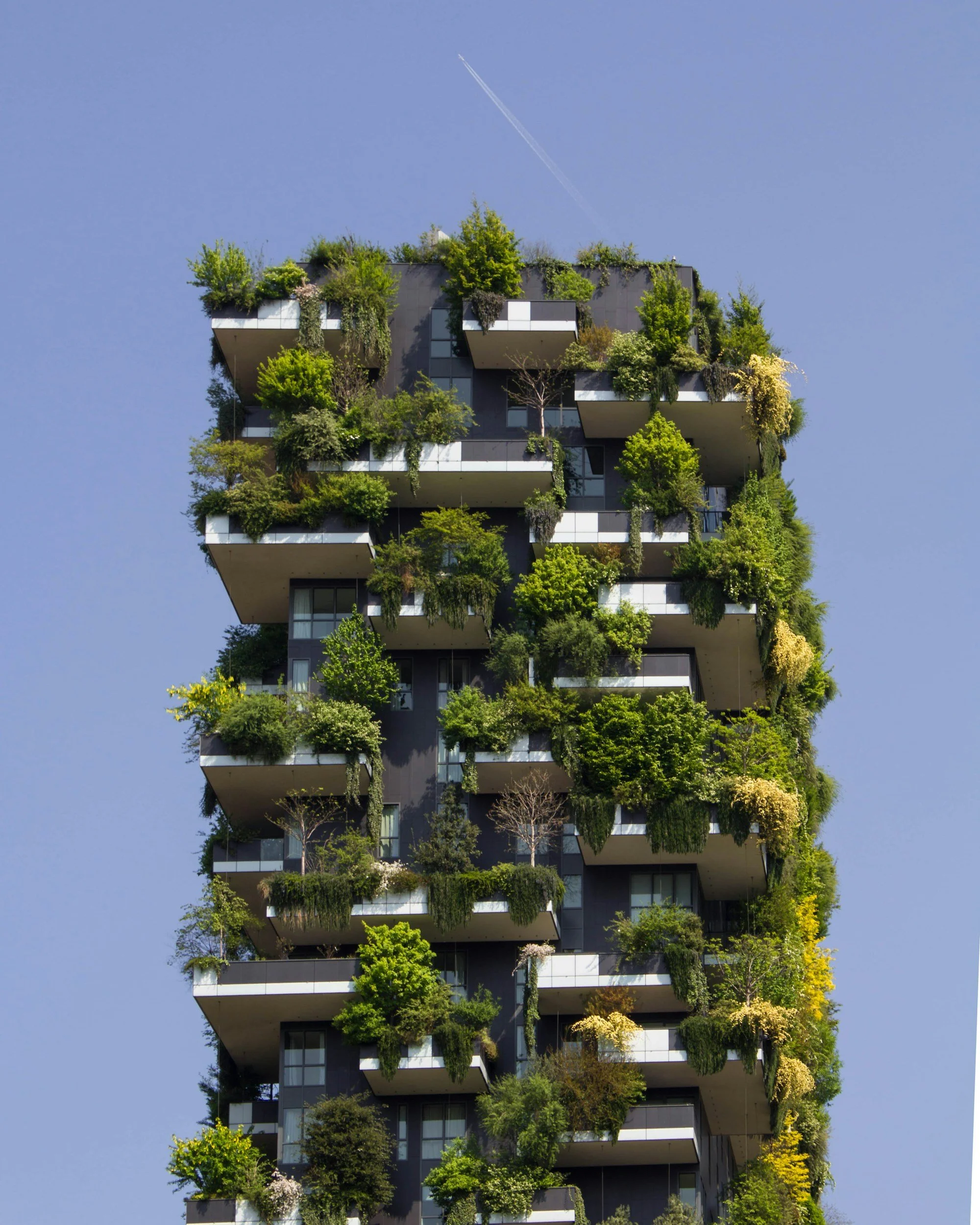 A tall modern building with numerous balconies filled with green plants and trees, set against a clear blue sky with a contrail.