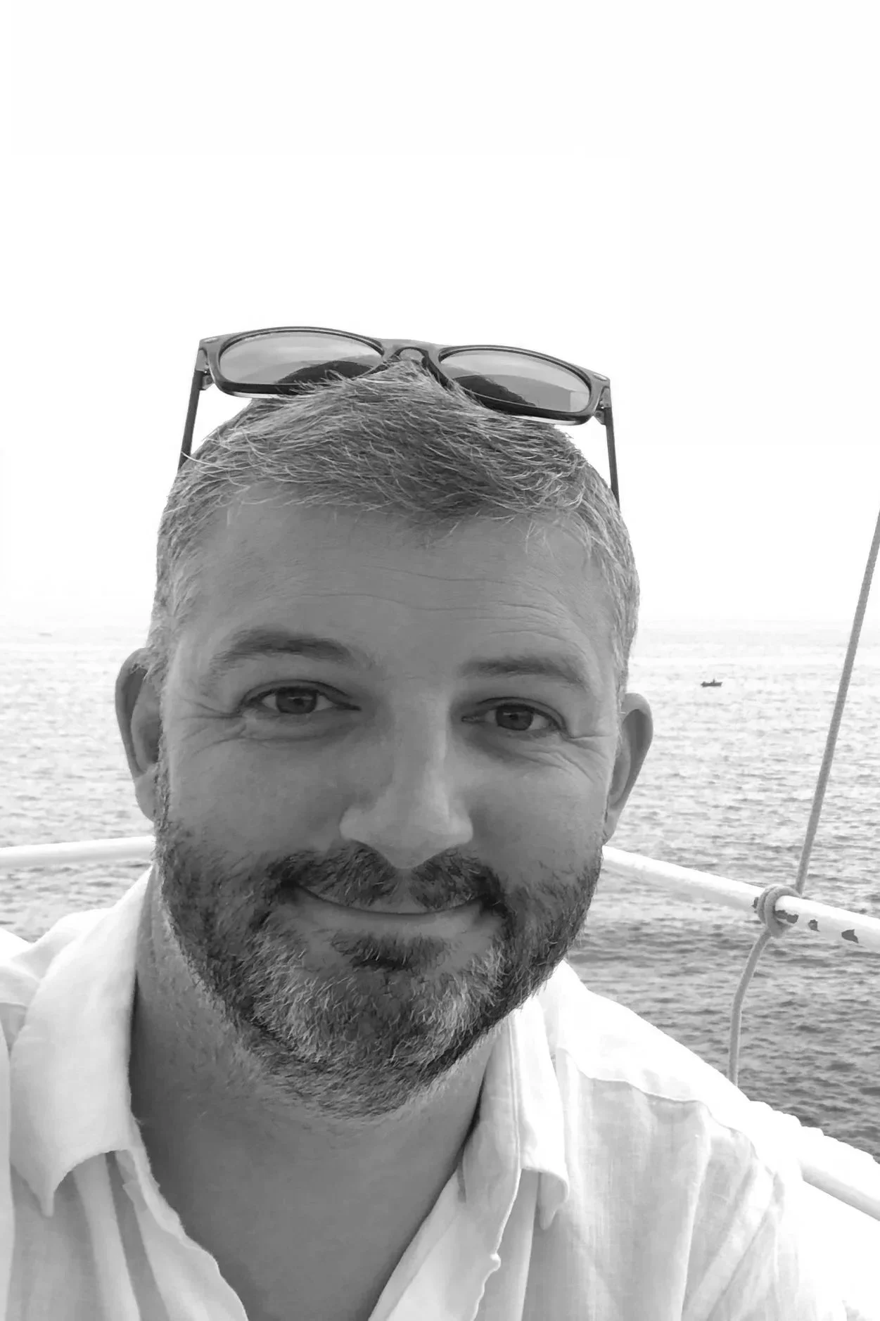 A smiling man with short hair and a beard, wearing glasses on his head, on a boat with the ocean in the background