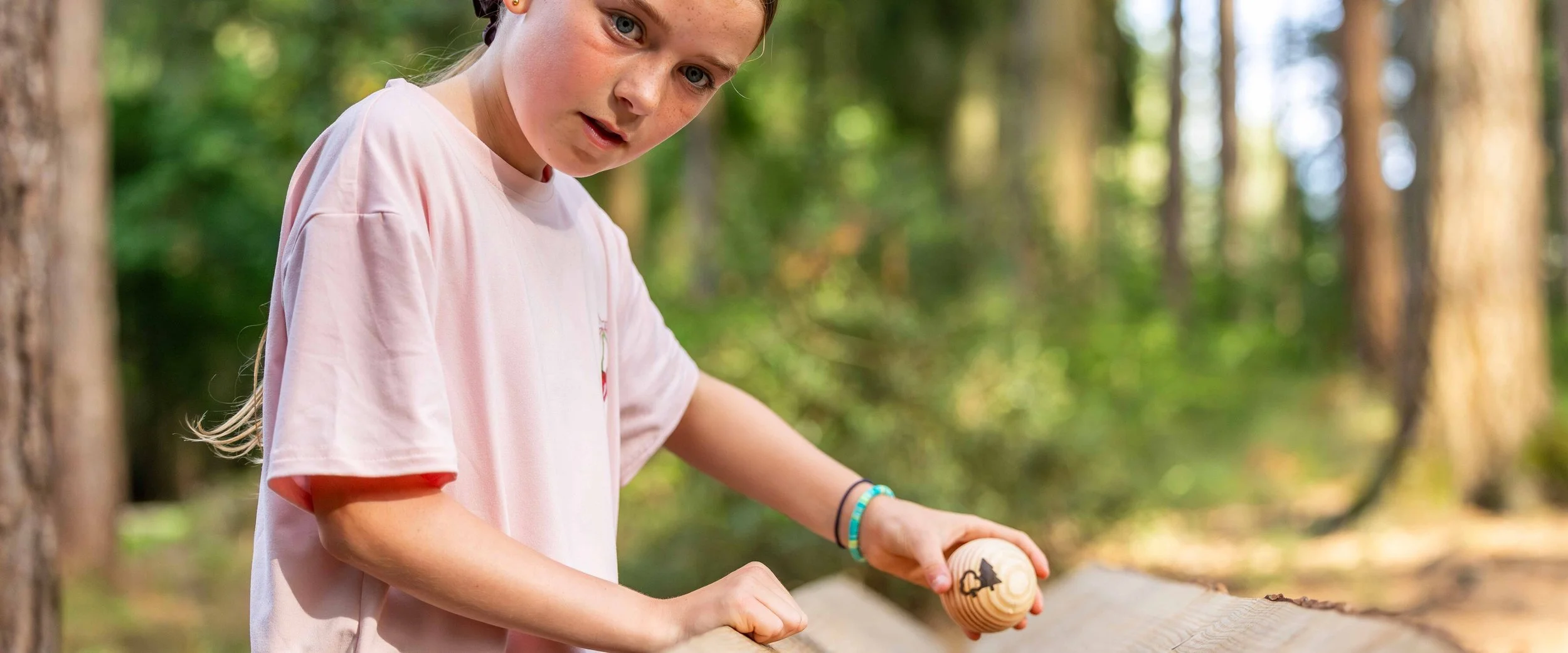 A young girl with light hair and freckles wearing a pink T-shirt, holding a wooden spinning top with a tree design, outdoors in a forest.