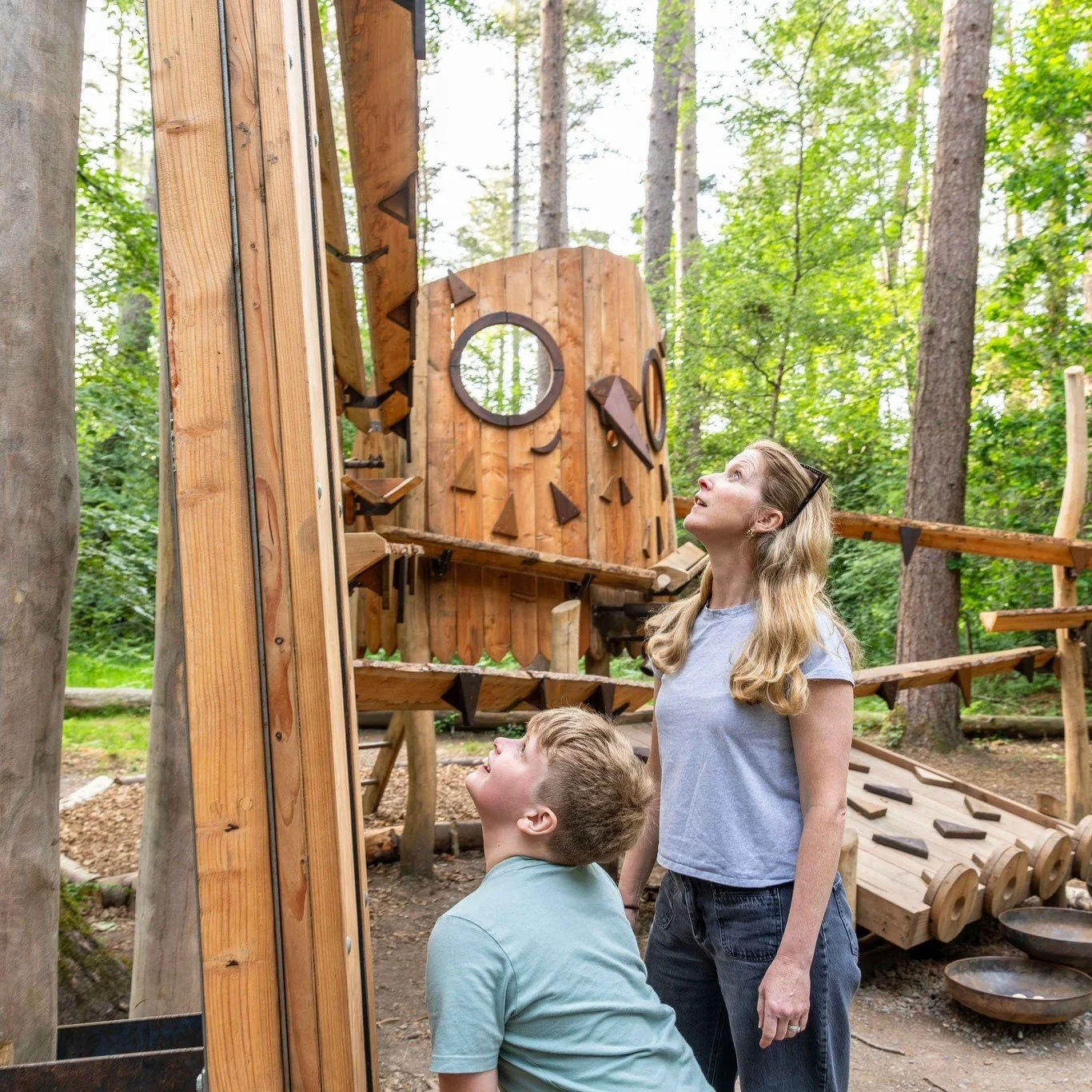 A woman and a boy looking up at a wooden sculpture in a forest.
