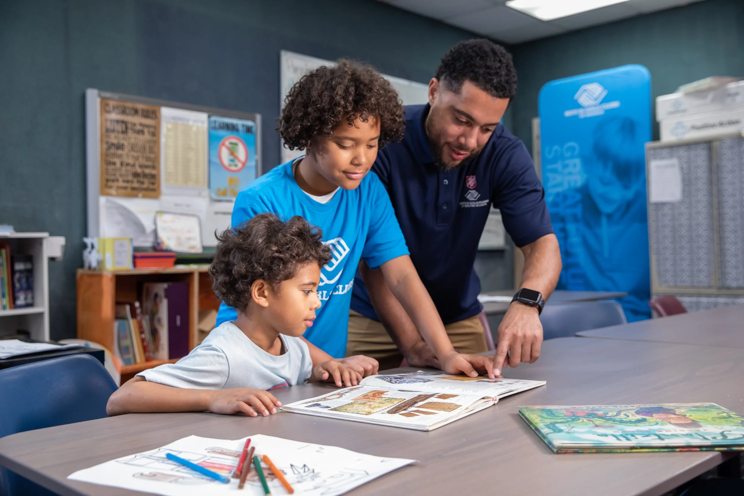 Two children and an adult reading a large book at a table in a classroom or library.